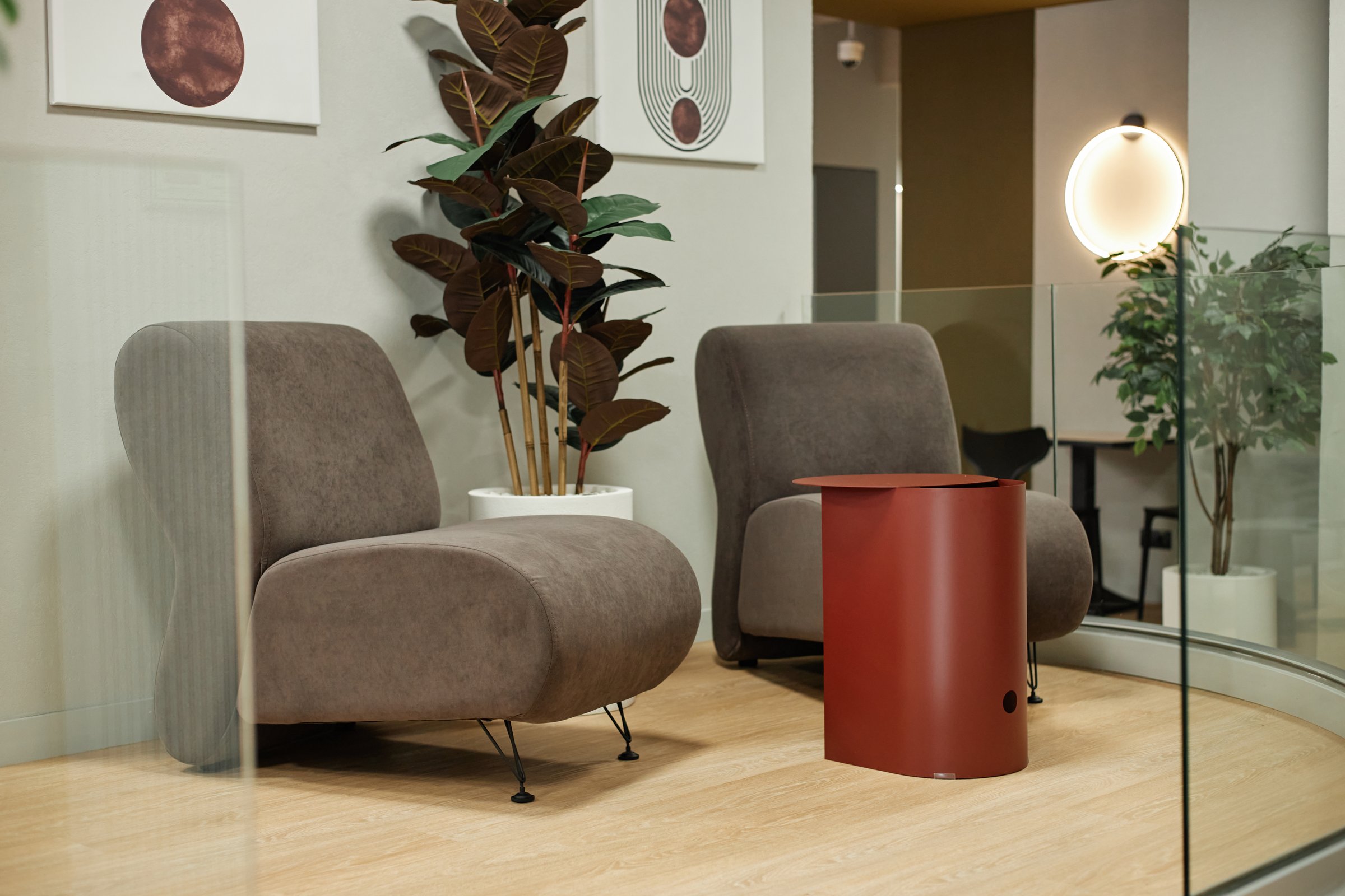 Modern waiting area featuring two empty armchairs and red table positioned on wooden floor with potted plants and abstract wall art