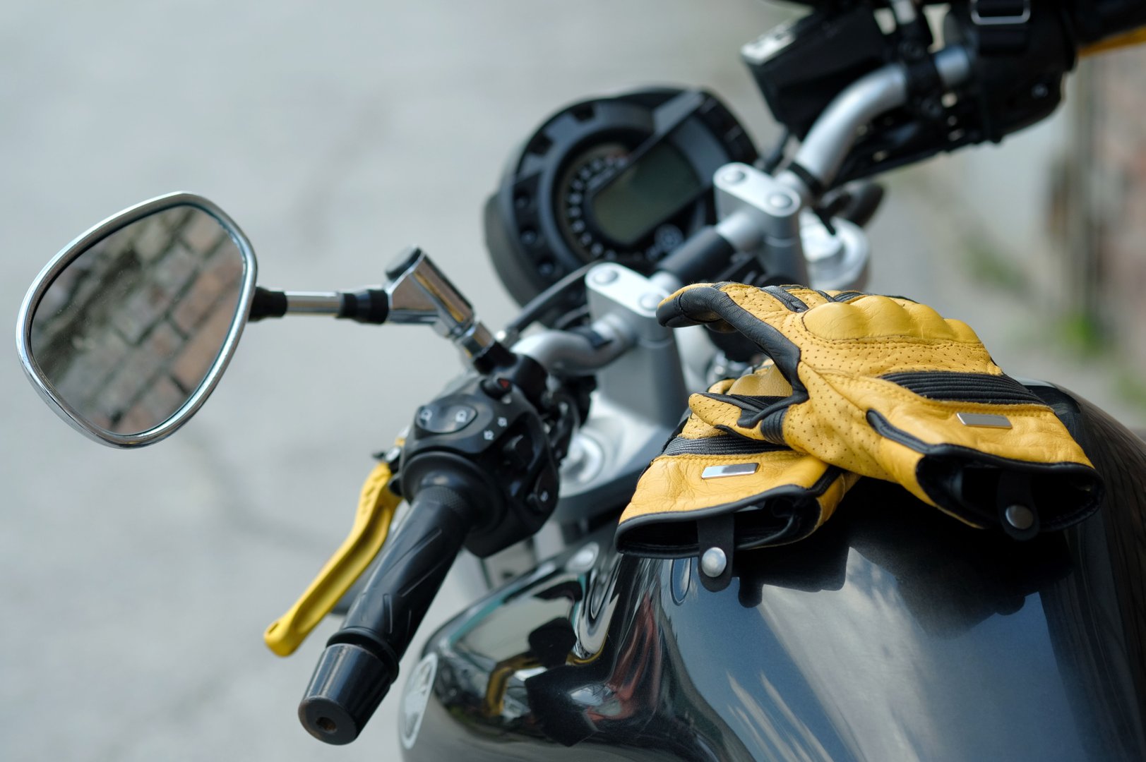 Yellow leather motorcycle gloves are on the black gas tank of a sports motorcycle.There are car control sensors in the background of the steering wheel.