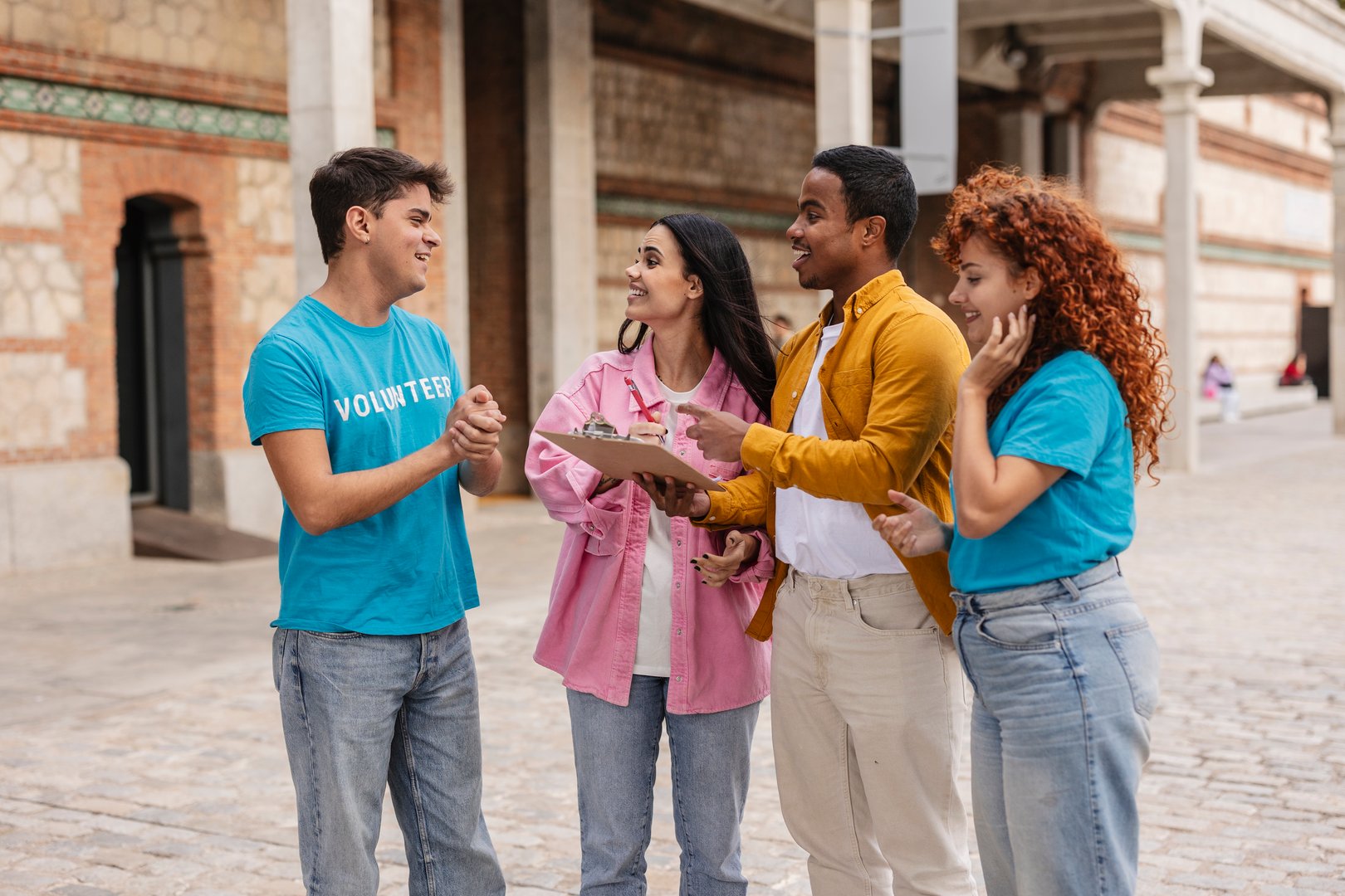 Four young adult volunteers wearing casual clothes are standing together outdoors in front of a brick building, discussing notes on a clipboard and planning their next steps during a charity event