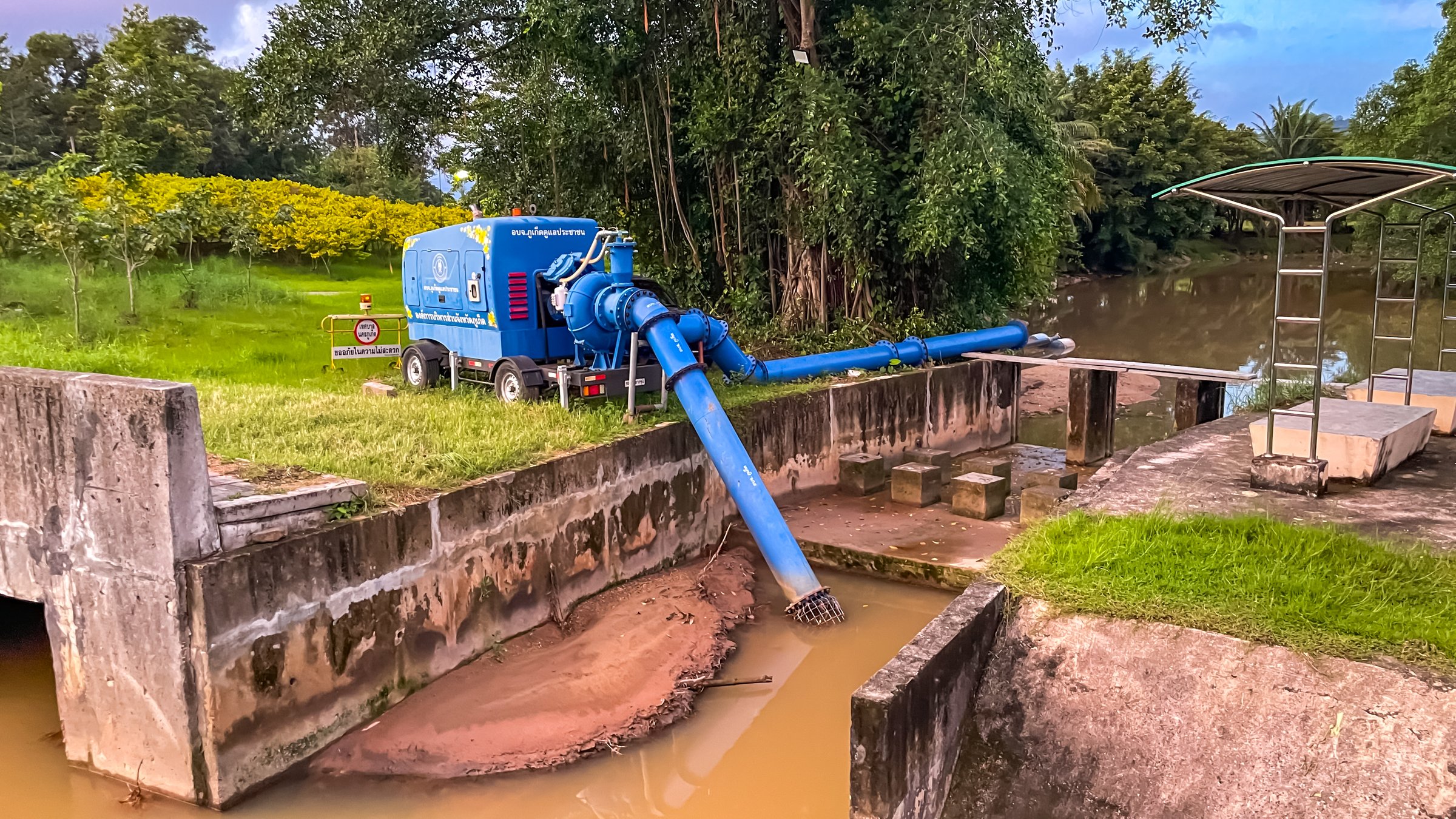Thailand, Phuket - November 1, 2025: A mobile pump with blue pipes pumps water from a canal; the equipment is installed on a concrete pad next to a river and green spaces.