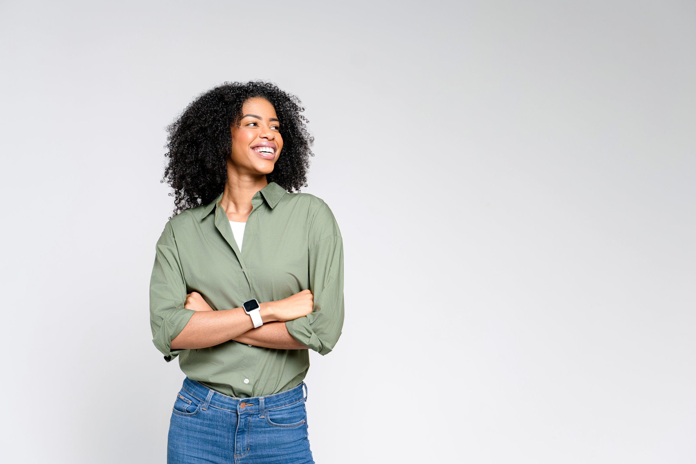 A confident African-American woman in a casual olive green shirt and jeans stands with a beaming smile, exuding a relaxed professionalism and easygoing charm against a light background.