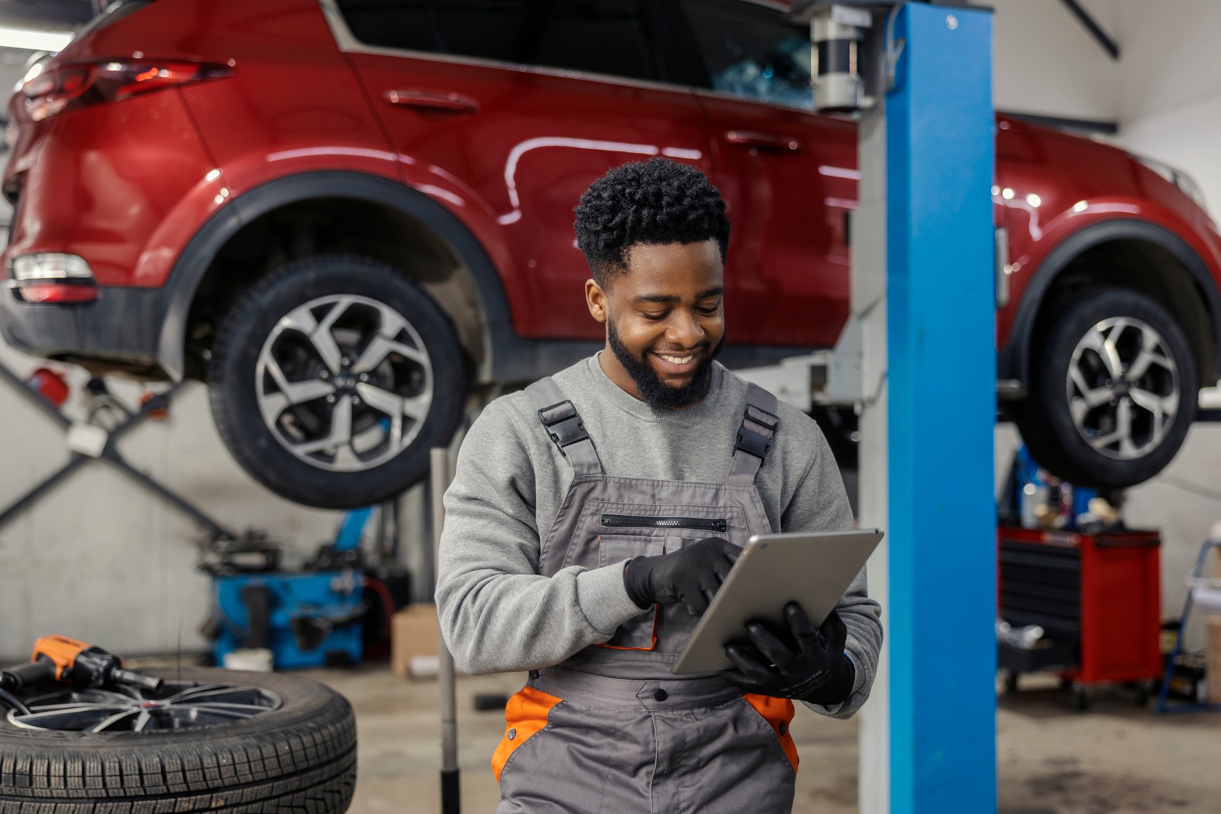 Smiling multicultural repairman standing at car service workshop with tablet in hands and scrolling on it.