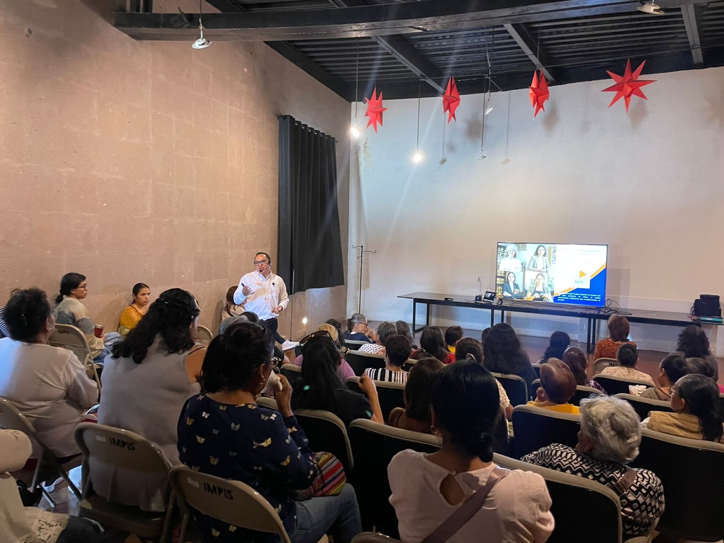 A group of people attending a presentation in a room with red star decorations and a screen showing images.