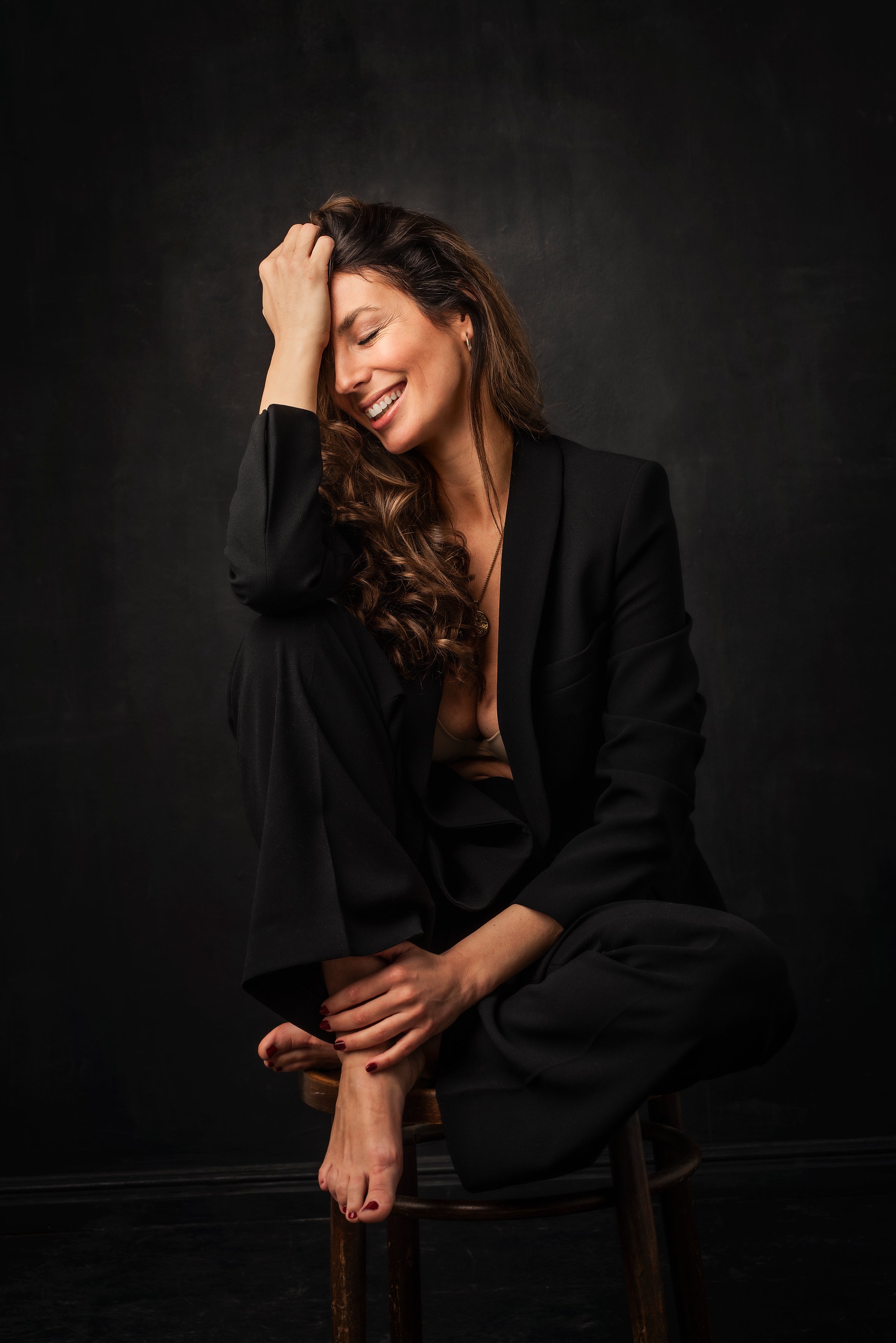 Full length of an attractive mid aged woman with toothy smile wearing black blazer and pants sitting against at isolated dark background. Copy space.