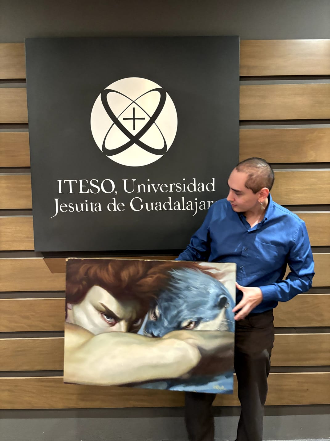 Man holding a painting depicting a human and a wolf. Background features an ITESO university sign on a wooden wall.
