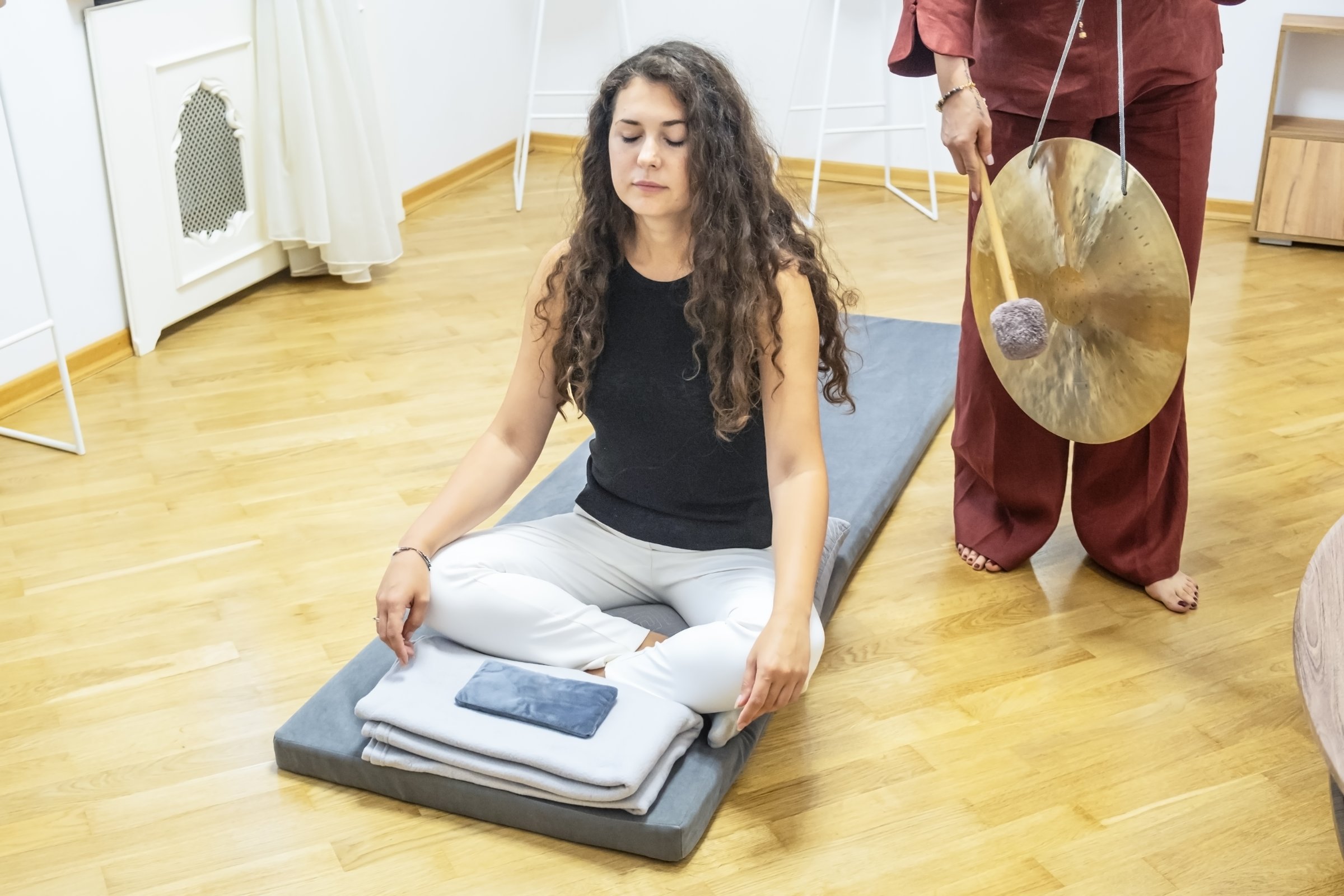 Young woman sitting in lotus position with closed eyes during a guided meditation session with gong sounds, practicing mindfulness and inner awareness in a wellness space