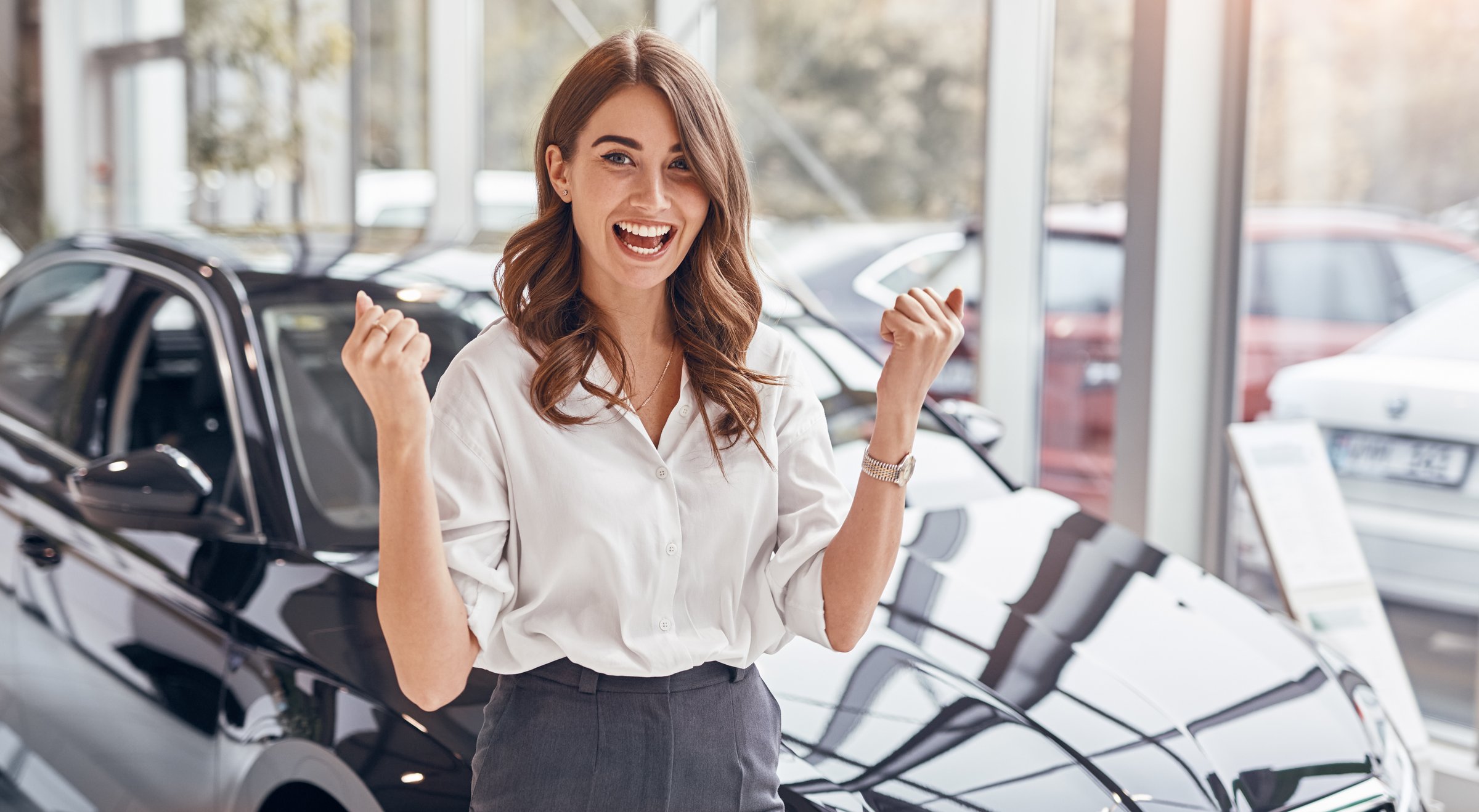 Cheerful female smiling for camera and clenching fists while standing near new vehicle in car dealership