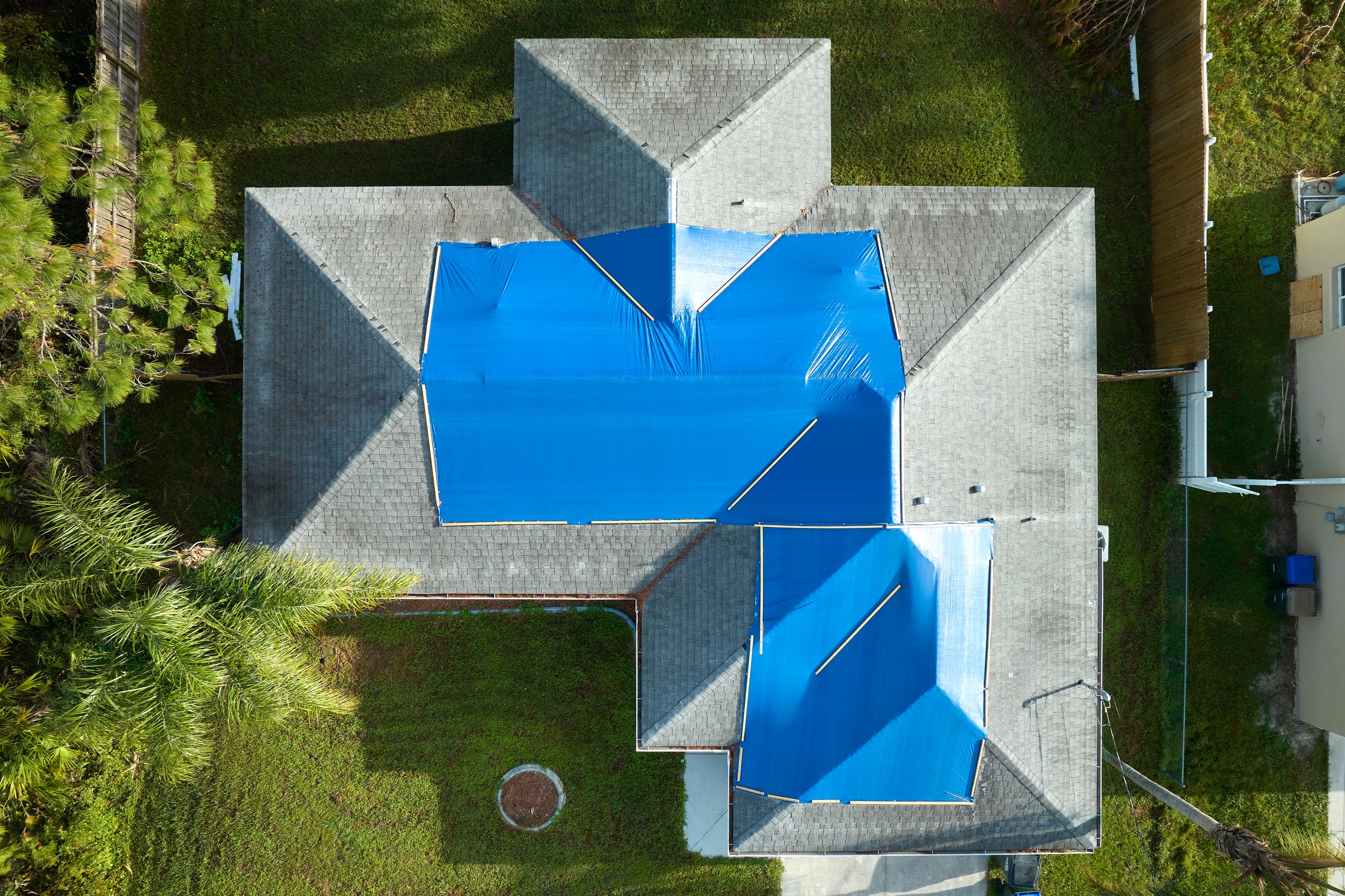 Aerial view of damaged in hurricane Ian house roof covered with blue protective tarp against rain water leaking until replacement of asphalt shingles.