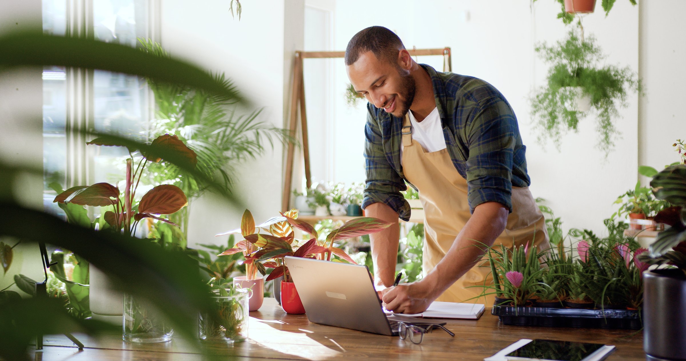African American man flower shop manager standing in apron in small floral shop. Businessman working in flower shop writing concentrated on business planning. Floristry business and retail concept.