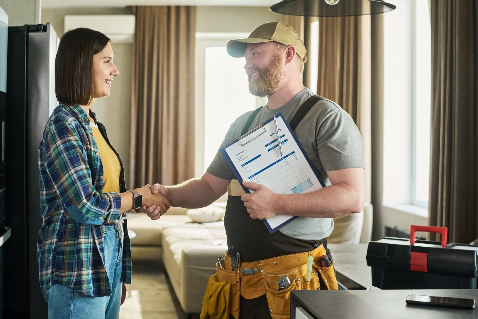 Caucasian young adult woman shaking hands with Caucasian middle aged male repairman holding clipboard and invoice, standing in modern living room, both smiling and making eye contact