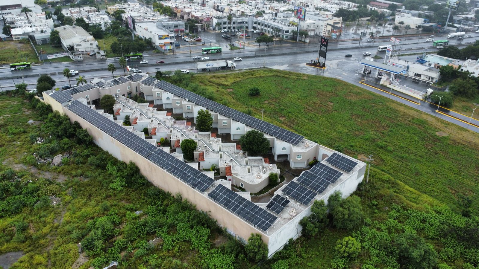 Aerial view of a row of houses with solar panels on a rectangular property, surrounded by greenery near a road.