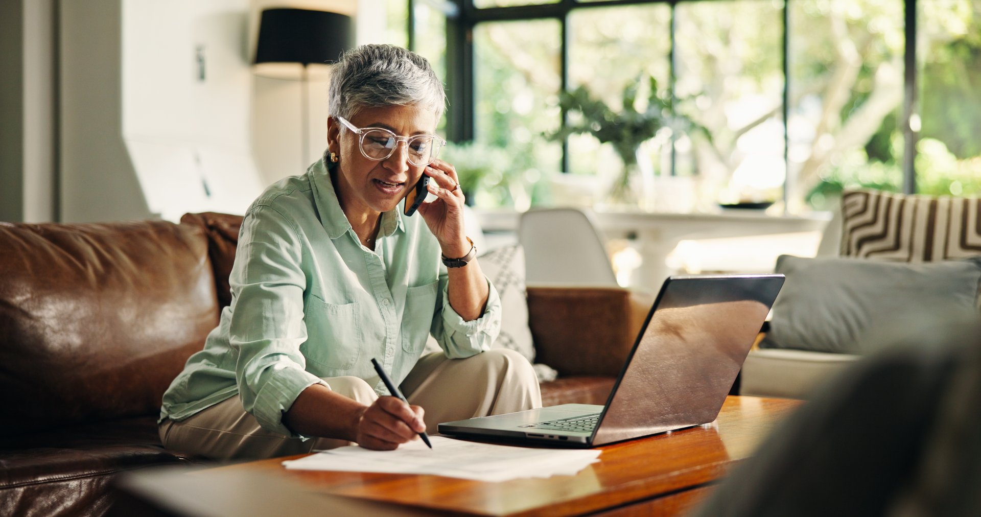 Phone call, laptop and mature woman writing notes for pension, planning and funds in home. Computer, cellphone and senior female person with discussion for investment document or account update