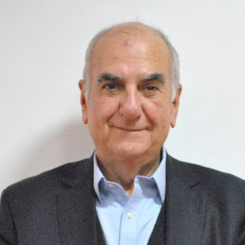 Elderly man with a slight smile wearing a dark suit and light blue shirt against a plain background.