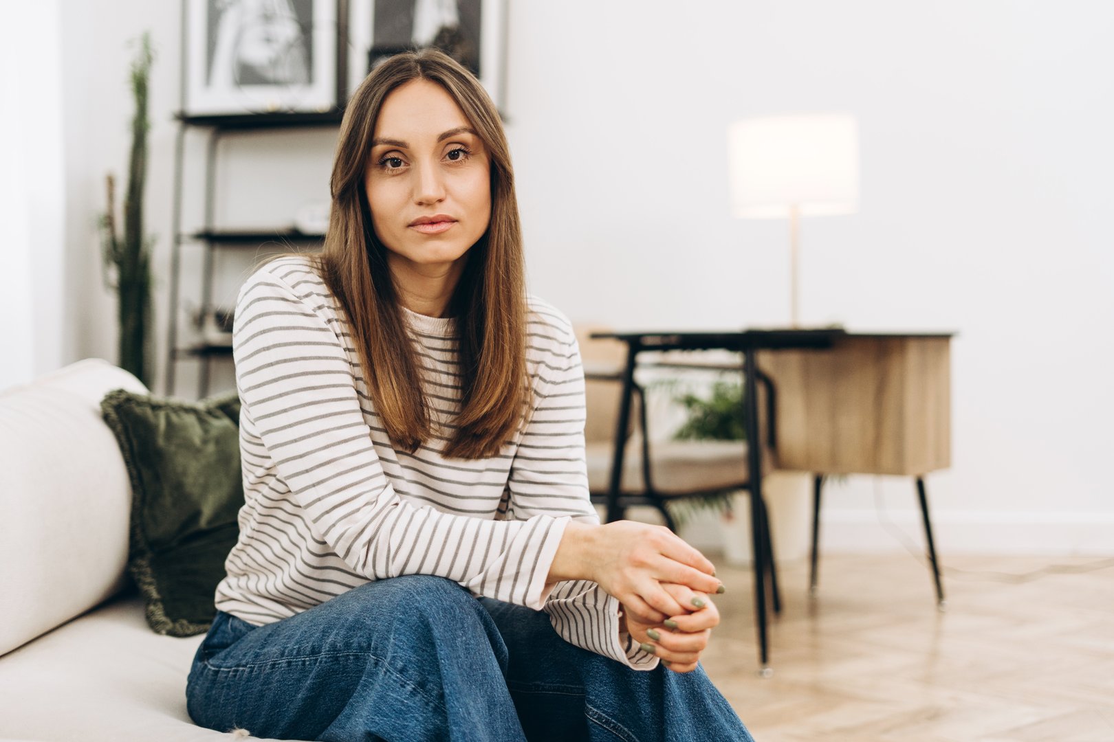 Young woman sitting on a comfortable sofa in a modern, stylish living room, lost in deep contemplation, hands gently clasped in her lap