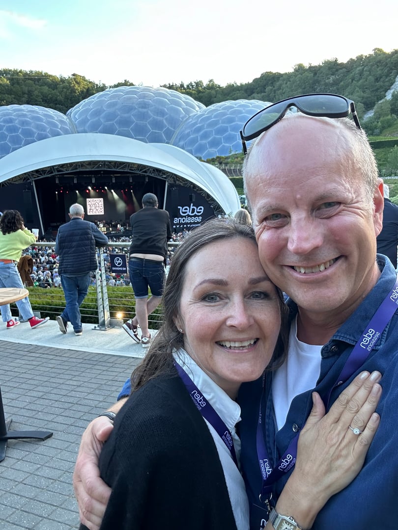 Two smiling people at an outdoor concert with a stage and audience in the background. They wear event lanyards.