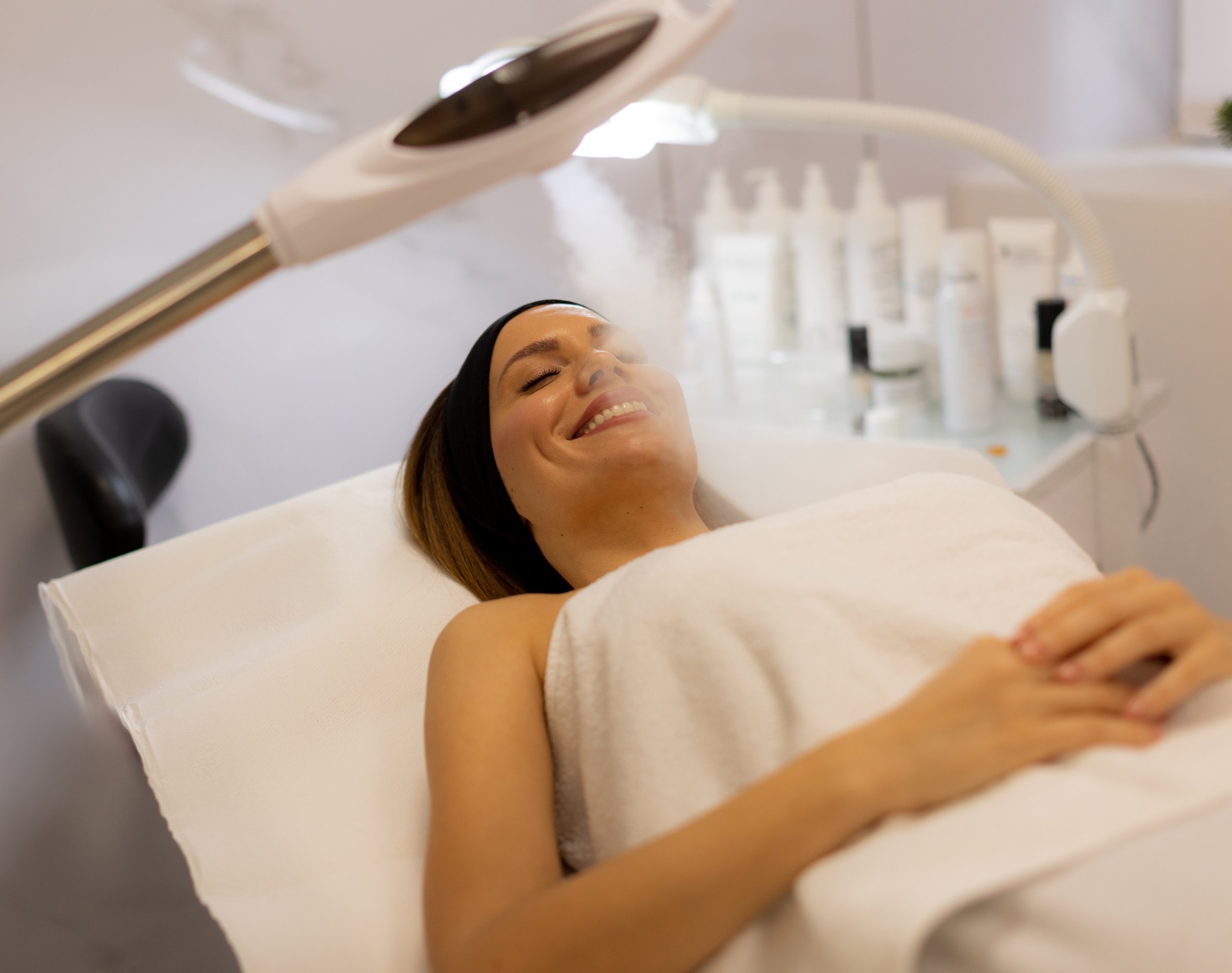 A tranquil spa setting features a woman enjoying a facial treatment, with mist enveloping her face. The atmosphere promotes relaxation and skincare rejuvenation.