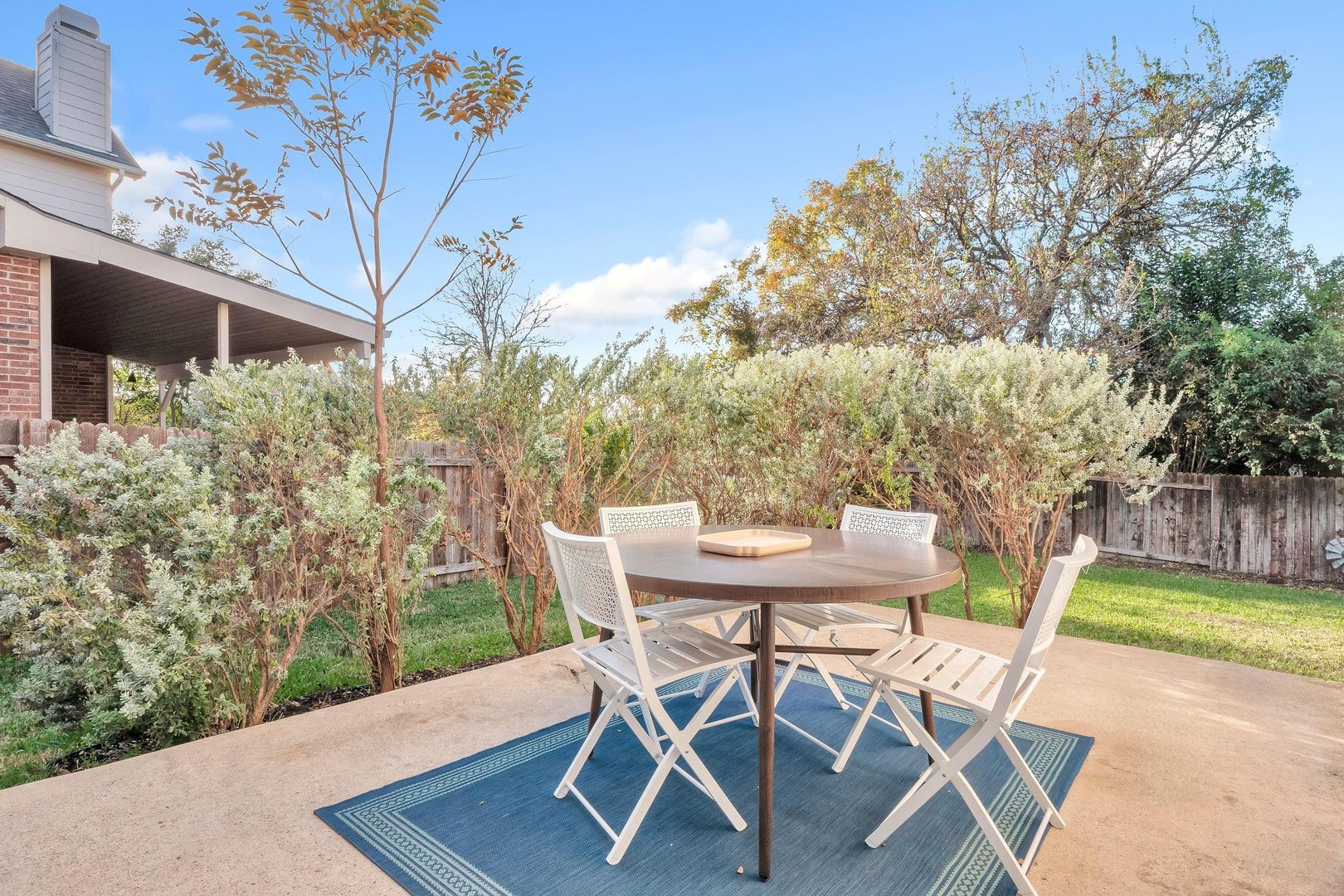Patio with round table, four white chairs on a blue rug, surrounded by greenery and a wooden fence.