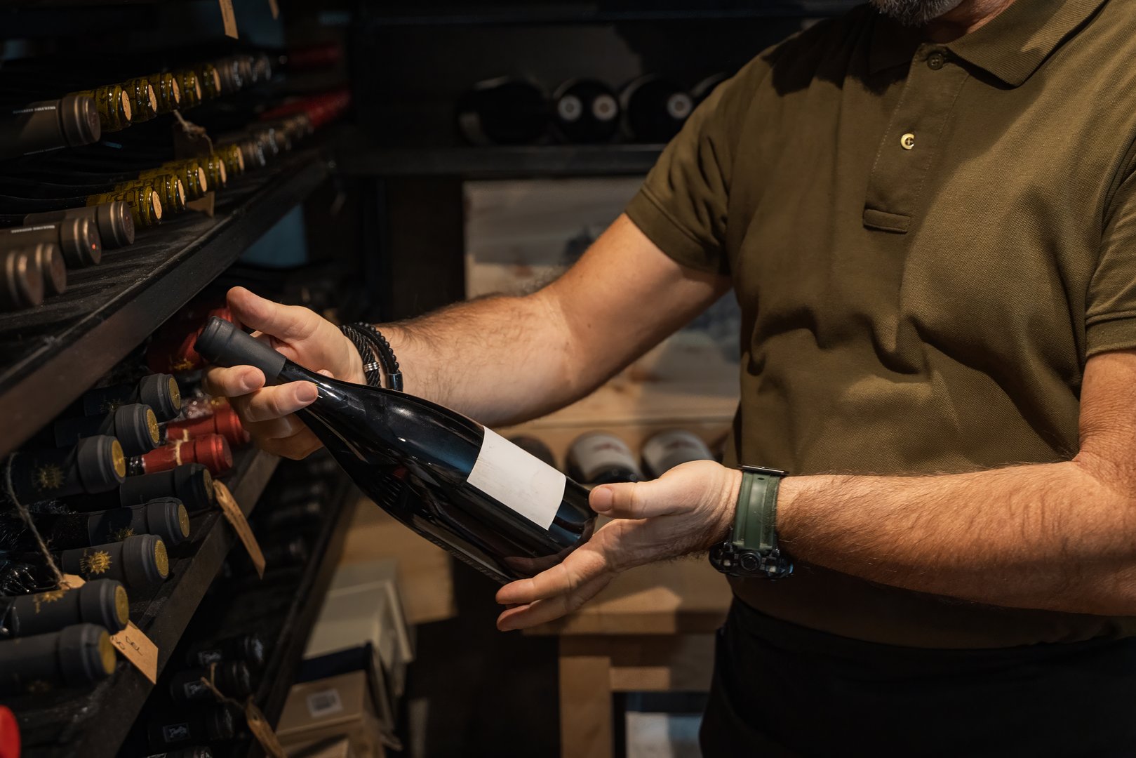Professional sommelier in casual uniform choosing wine bottle from wooden rack in dimly lit cellar. Luxury wine storage and collection concept with focus on hands