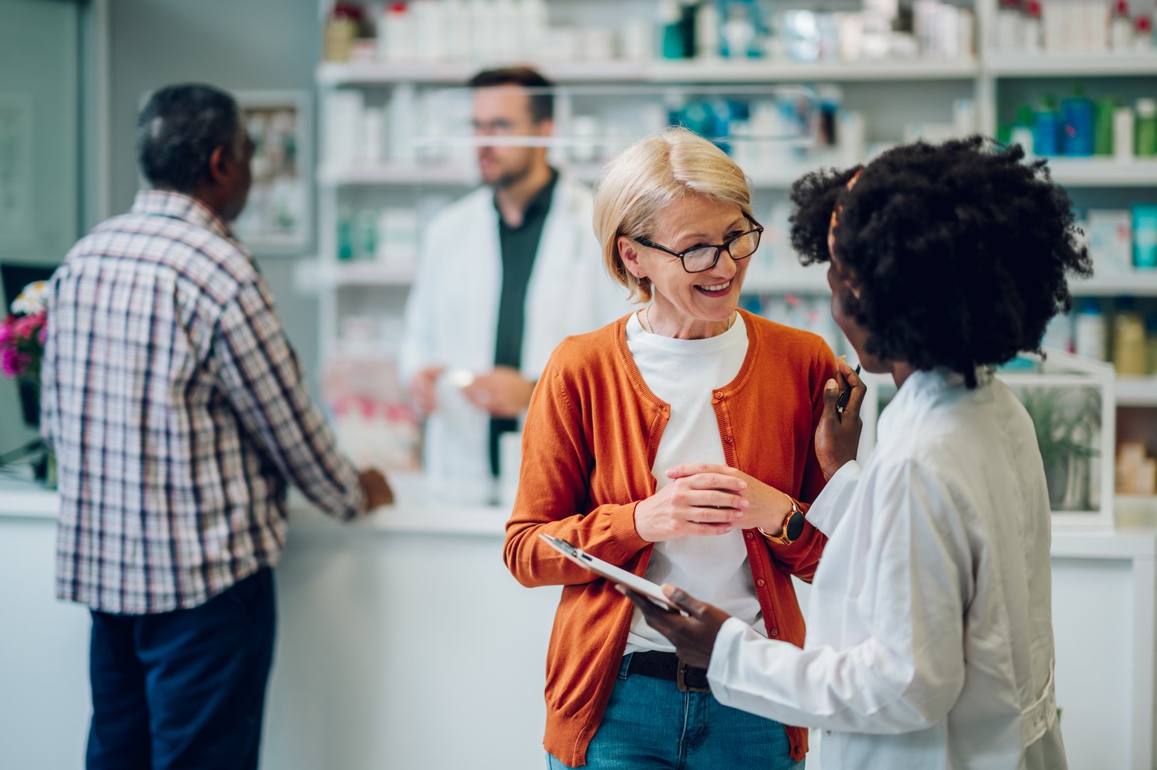 Young black woman pharmacist advising senior female customer about medications in a pharmacy. Shelves with health care products and a male colleague pharmacist in the background. Copy space.