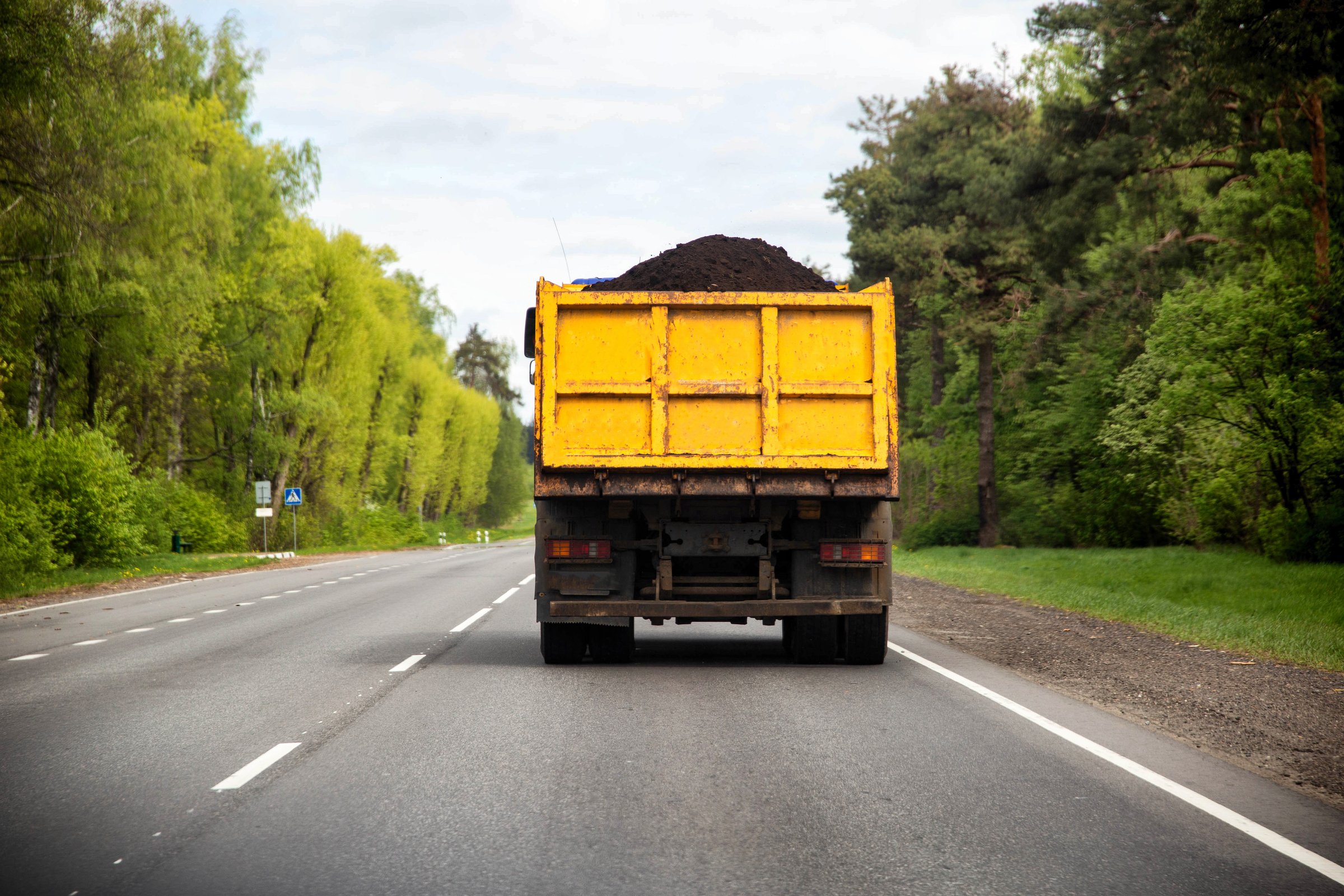 A dump truck with a yellow body transports black soil along an asphalt road against the backdrop of a forest