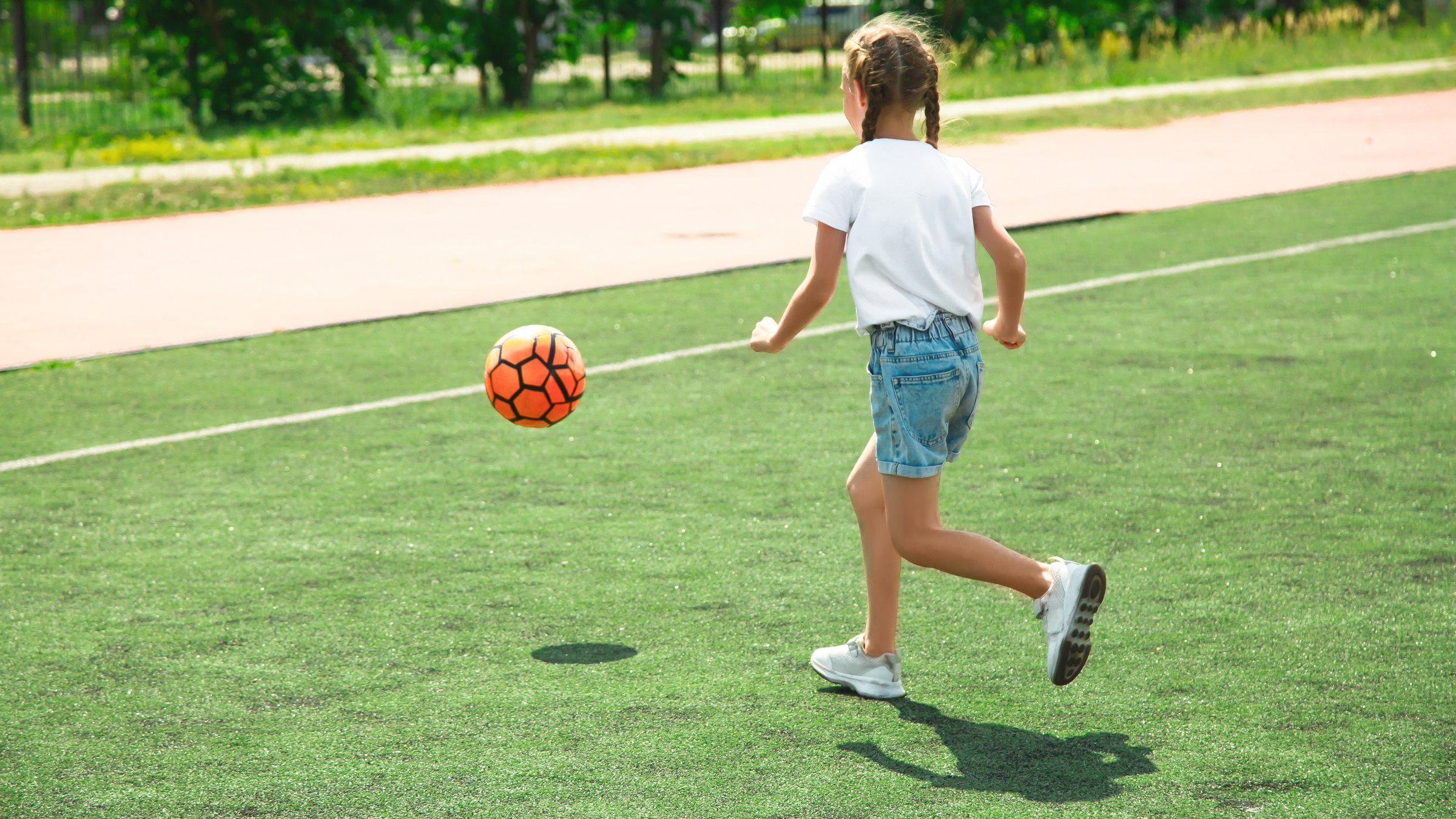 childhood, leisure games and people concept - happy little girl with ball playing soccer at school field