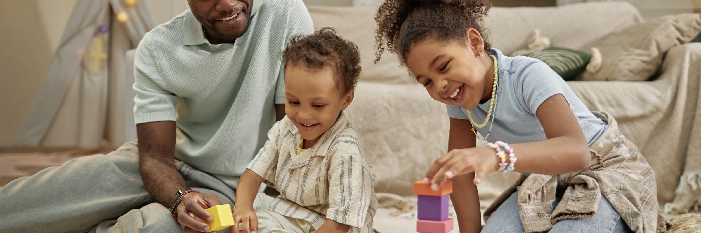 Website header shot of smiling African American children building constructions with toy blocks playing creative game together with father, while sitting on floor in cozy living room