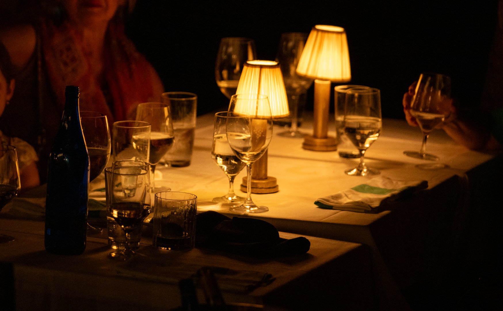 Sets a welcoming atmosphere for an evening gathering featuring elegantly set tables with warm, soft lighting. Glasses of different shapes and sizes reflect the gentle glow, suggesting conversations, laughter, and a shared meal among friends.