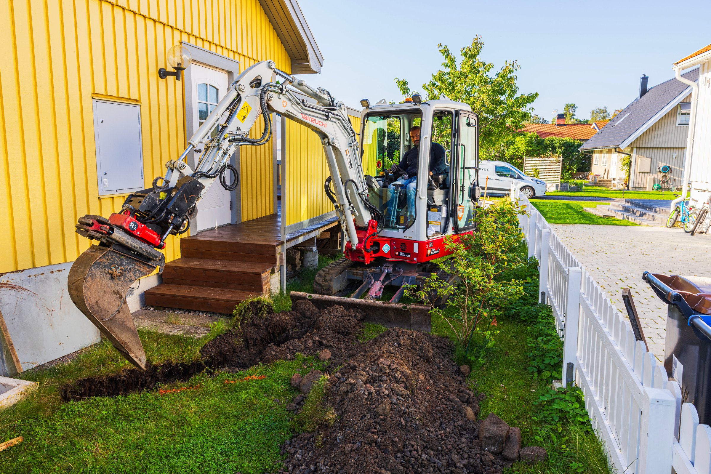 Sweden. Stockholm. September.10.2025. Takeuchi TB225 compact excavator operated by worker digging trench near yellow wooden house with garden and fence.