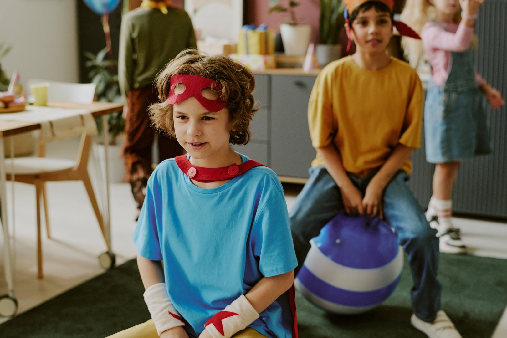 Caucasian boy wearing superhero costume sitting in foreground, other children playing during indoor birthday party