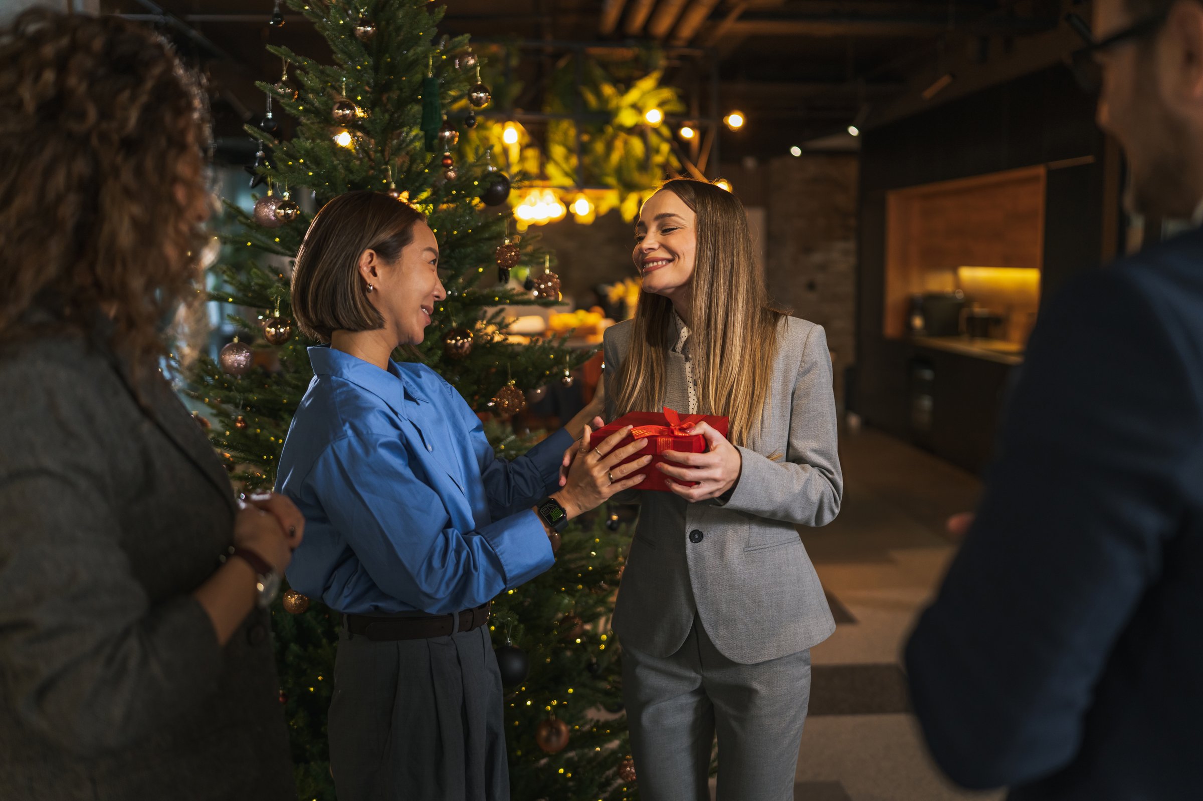 Female colleague giving a red present box to another woman with a surprised expression during an office christmas party, enhancing team spirit and holiday celebration
