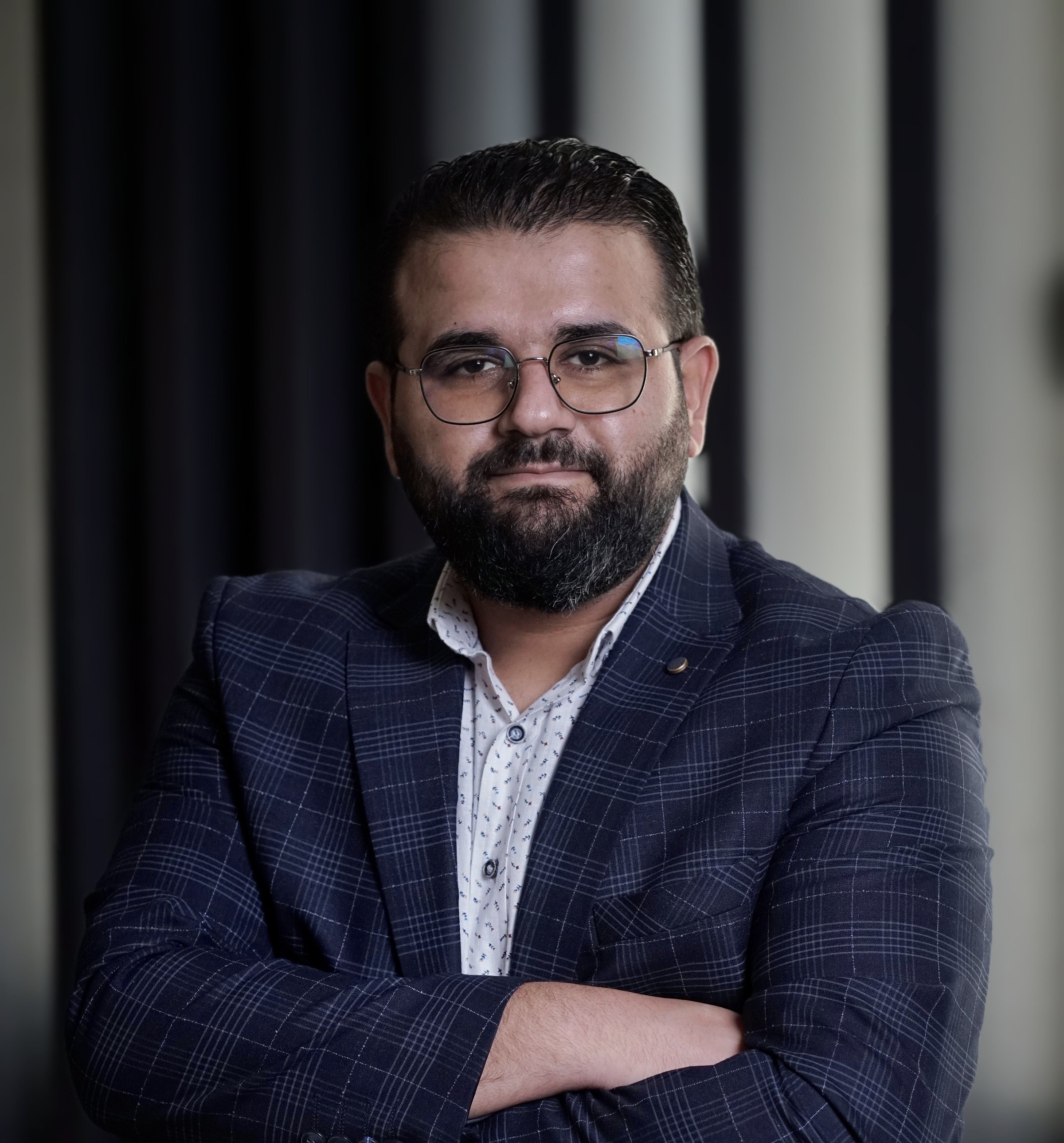 Man with glasses and beard wearing a checkered suit jacket and white shirt, standing with arms crossed.