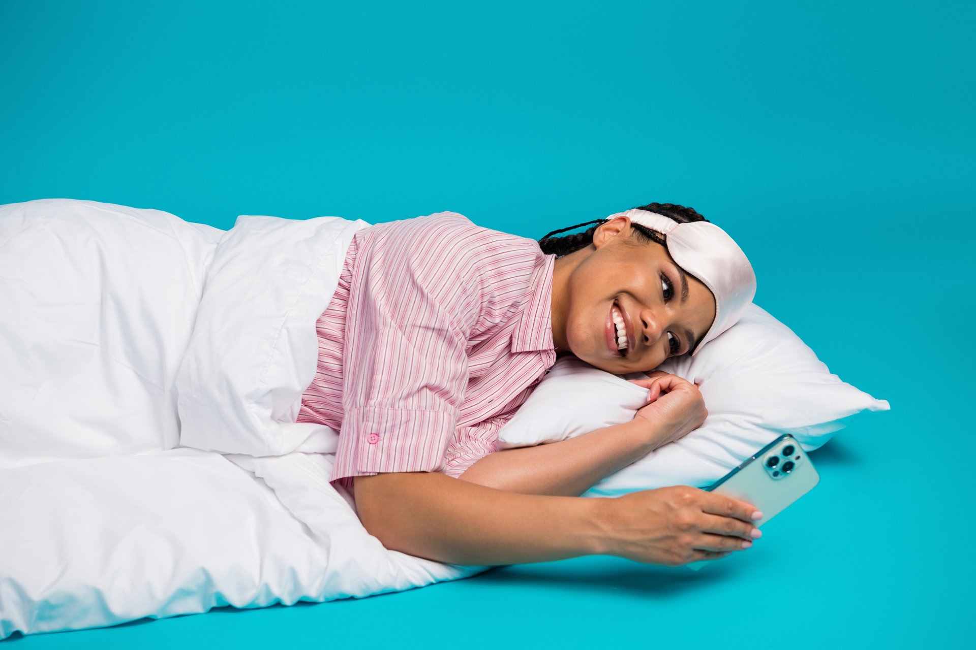 A cheerful young woman in pink-striped pajamas lies in bed against a cyan backdrop, smiling warmly while holding her smartphone.