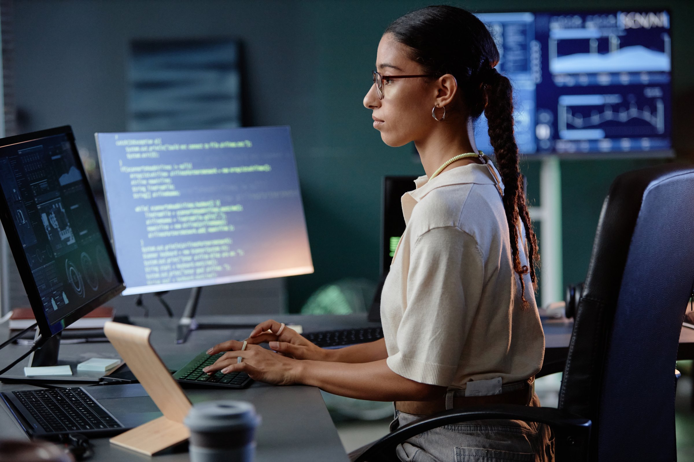 Side view of confident Middle Eastern female programmer typing on computer keyboard debugging code while working in evening hours at desk in IT company office