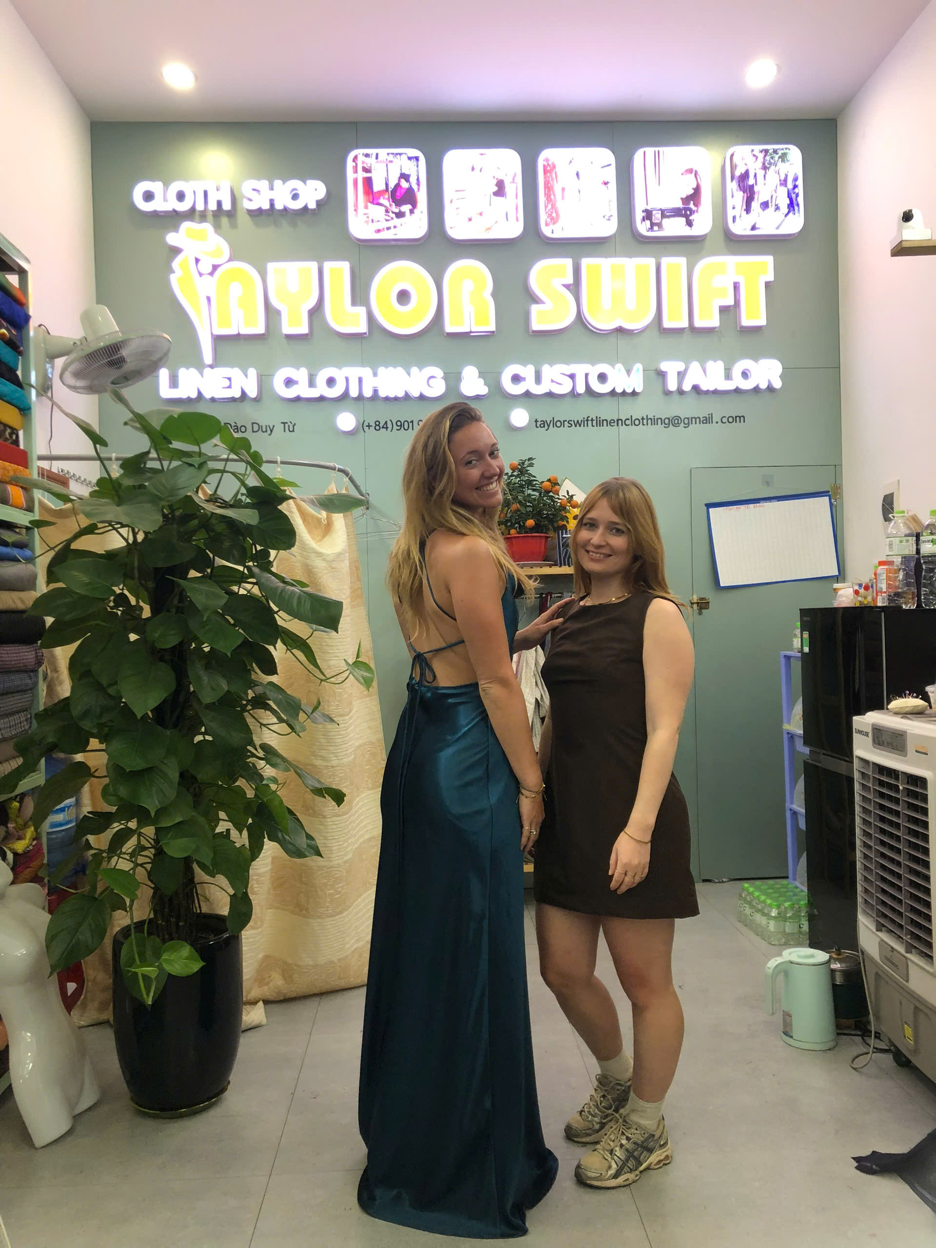 Two women pose in a clothing shop with a "Taylor Swift" sign, surrounded by fabric and tailor equipment.
