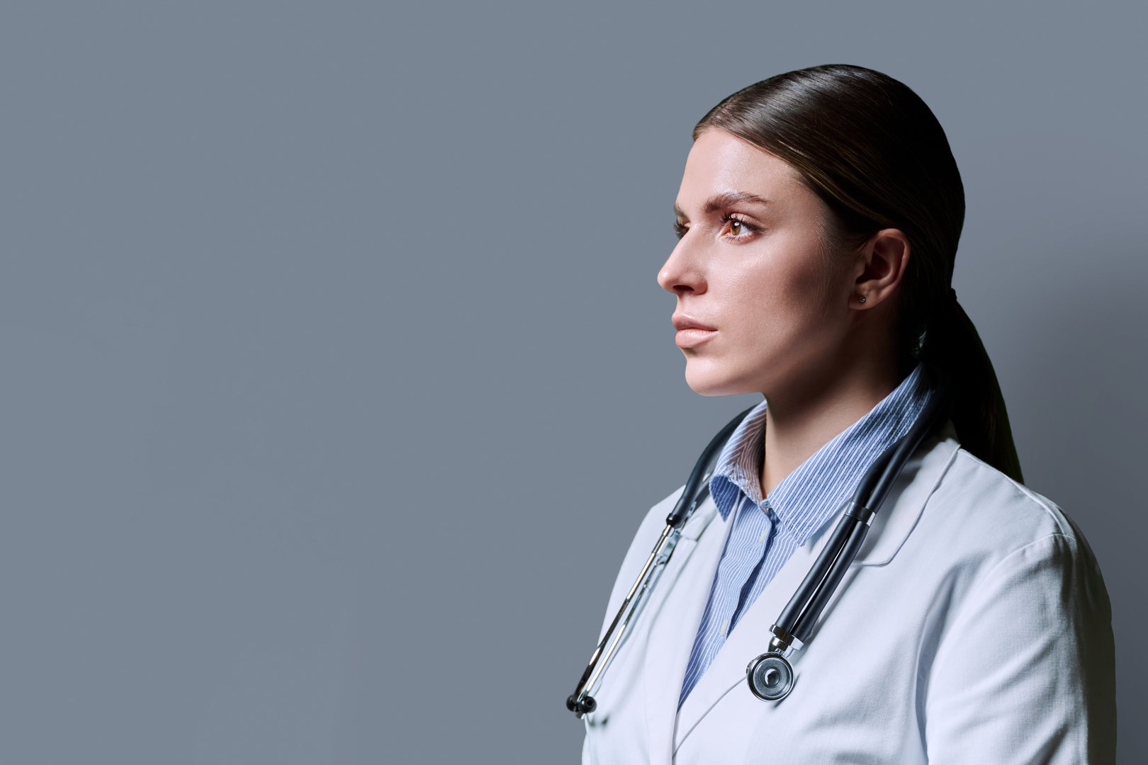 Profile view of young confident serious female doctor with stethoscope on gray studio background, copy space. Medicine, healthcare, treatment, medical staff concept