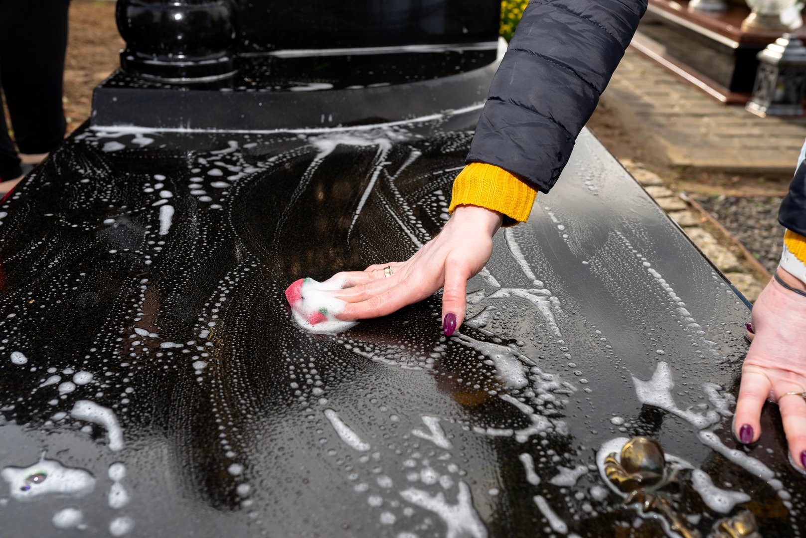 Cleaning a black tombstone with a cloth and water, female hands visible.
