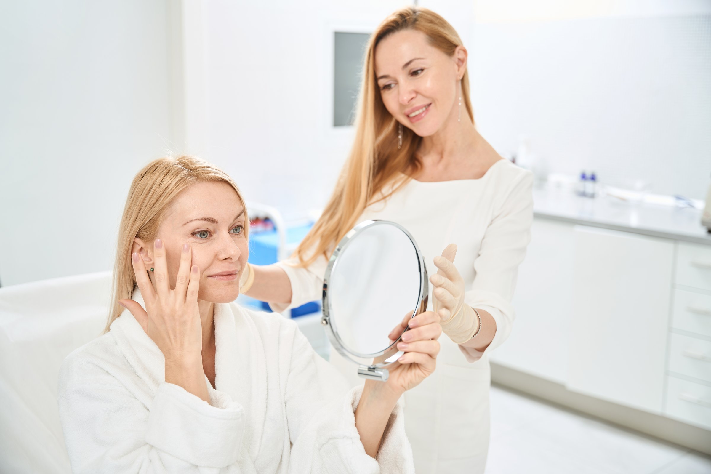 Smiling woman client of aesthetic medicine clinic satisfied with scar removing and skin lifting around eyes, looking at her reflection in the mirror, good service