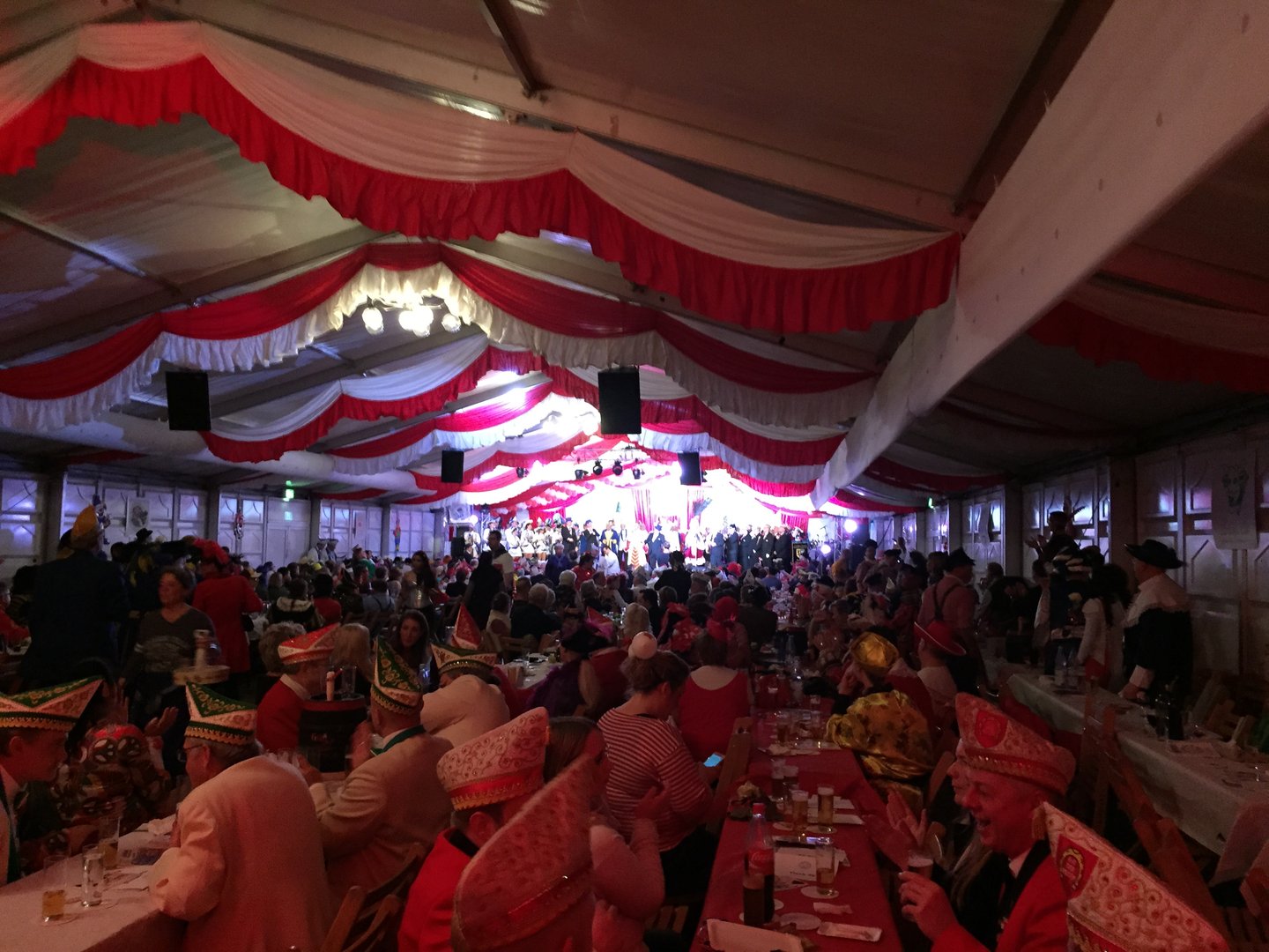 People in costume and colorful hats gathered in a large, festively decorated tent with red and white drapes.