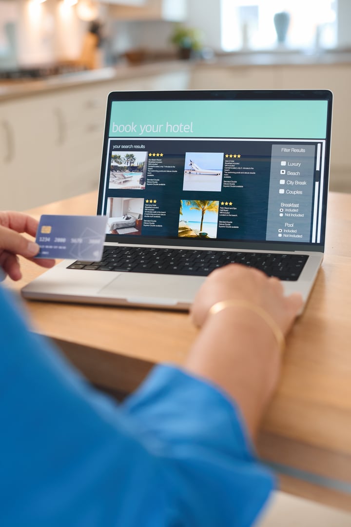 A person is using a laptop to book a hotel online while holding a credit card in their hand. The screen displays various hotel options with ratings and pictures, set against the backdrop of a contemporary kitchen.