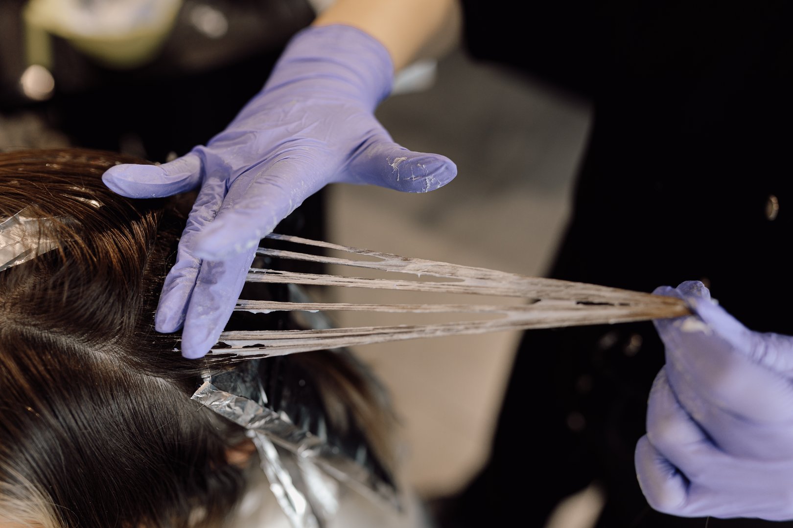 Top view of woman sit in beauty salon. Unrecognizable woman hairdresser hairstylist colorist hold strand of long hair, distributing hair dye with fingers in blue rubber gloves. Highlighting, balayage.