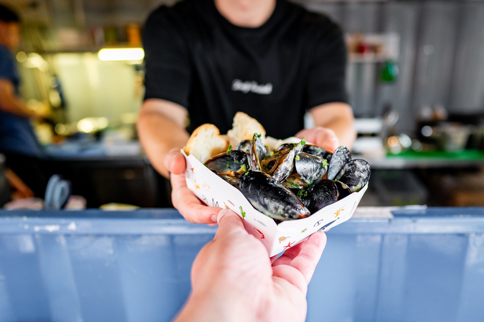 Close-up of hands offering a paper tray of cooked mussels, with a blurred kitchen background