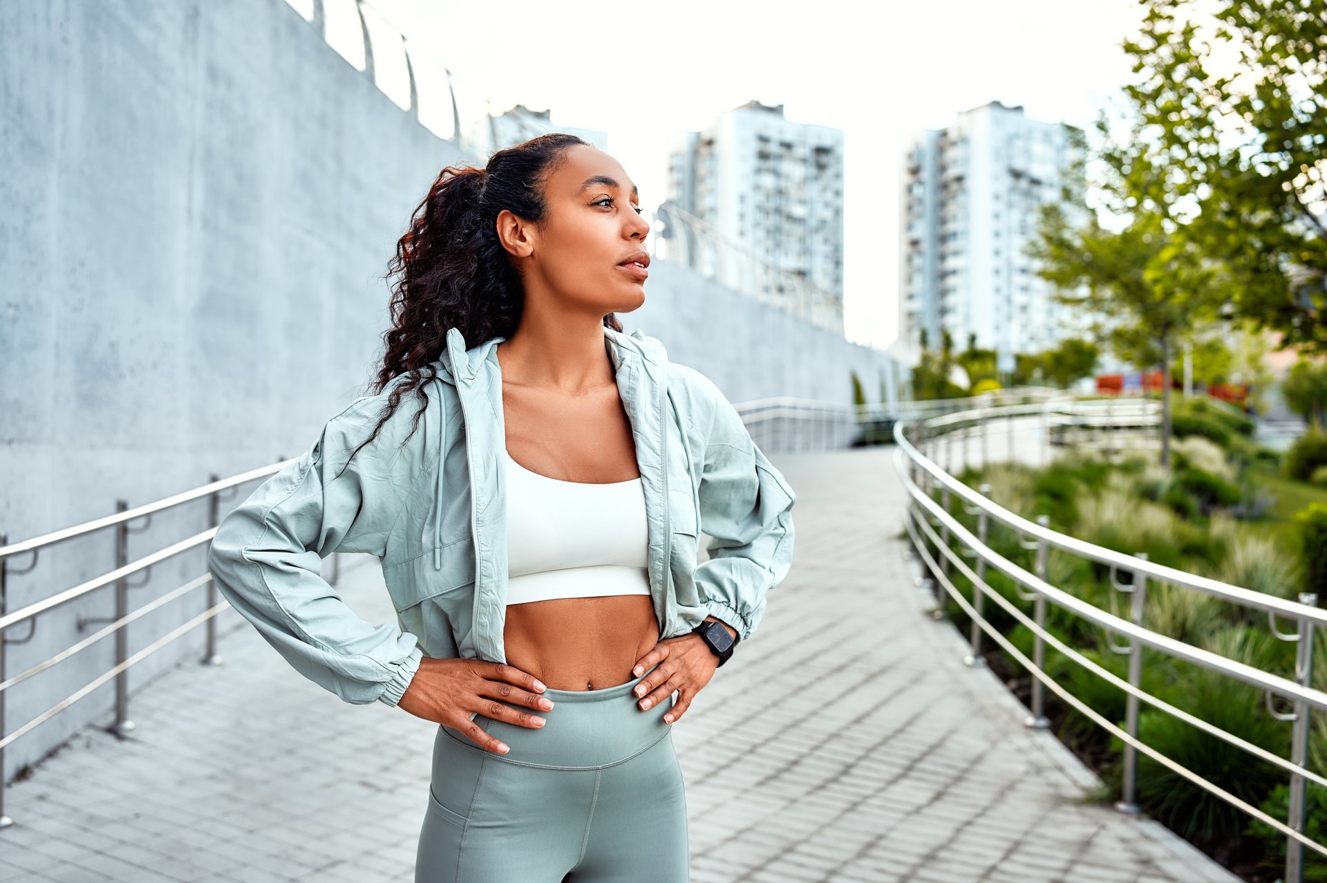 A beautiful sportswoman in a white top and soft blue jacket and sports leggings stands on a jogging track in the park outdoors and looks away. Preparation for a morning run.