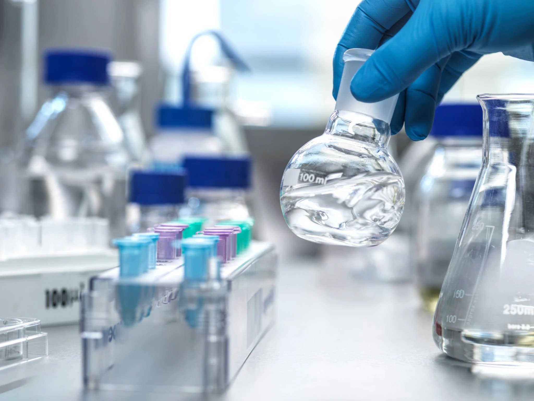 A gloved hand holds a flask filled with a clear liquid in a laboratory setting, ready for scientific analysis. Other equipment is in the background.