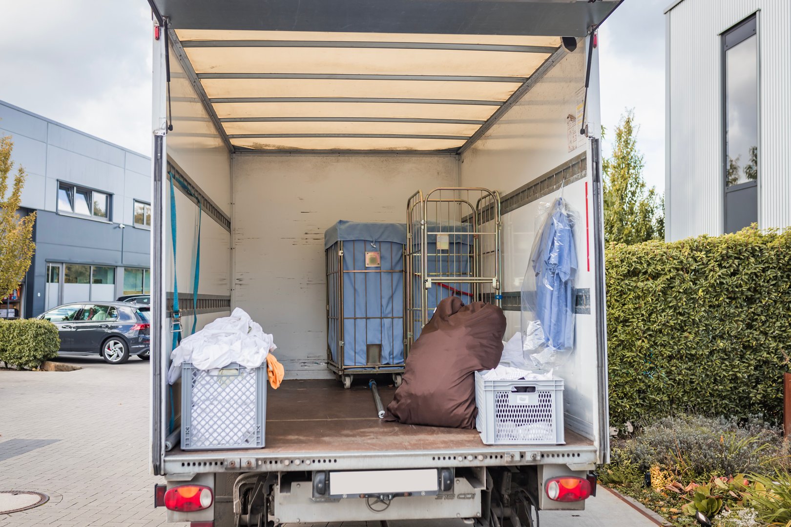 An open moving truck is pictured in an urban area, loaded with crates and an assortment of boxes. The setting conveys a busy, commercial atmosphere with surrounding buildings.