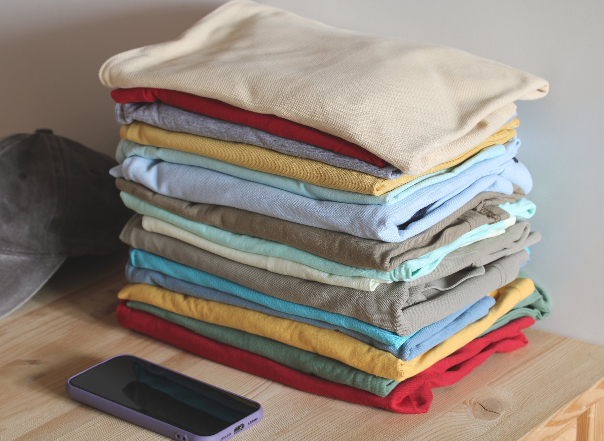 Many folded colorful t-shirts are stacked on a wooden table next to a smartphone and a cap
