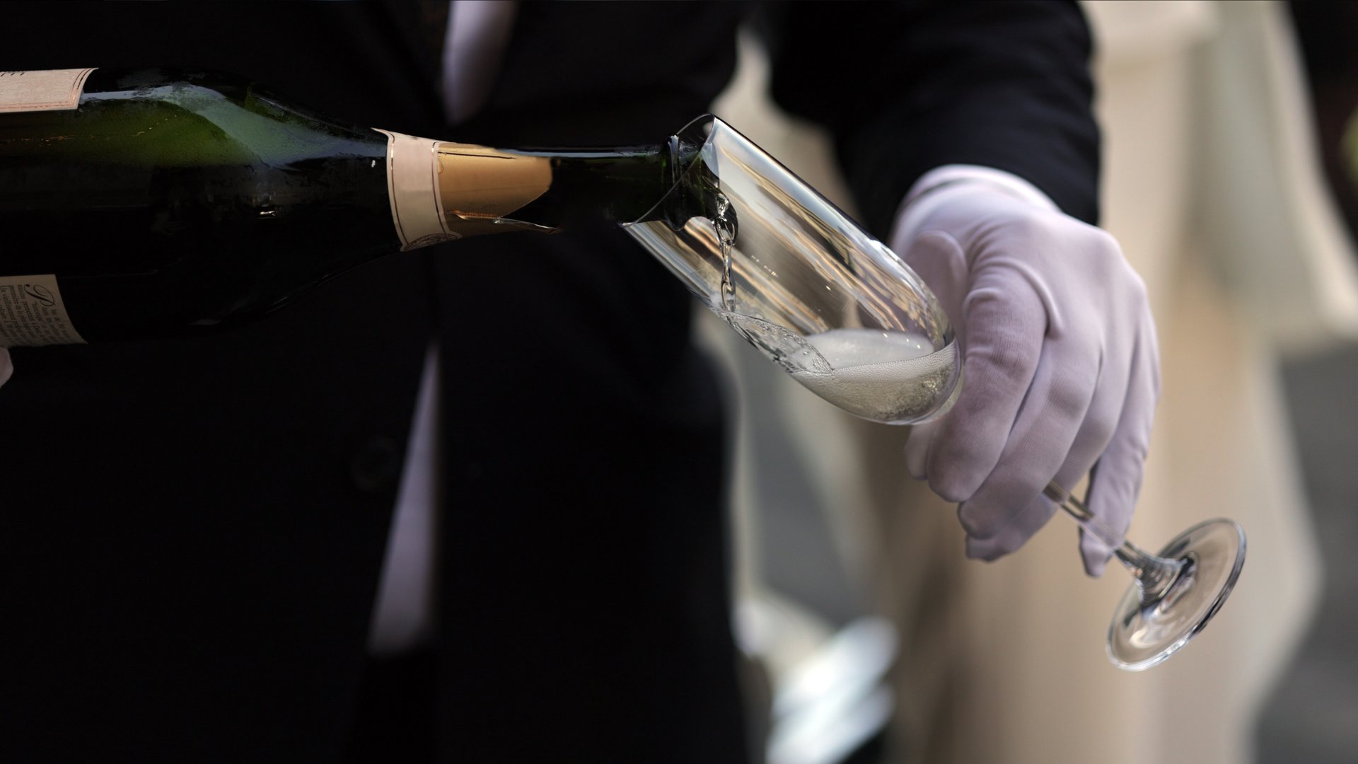 A close-up of a professional server wearing white gloves while pouring champagne into a slender flute glass, highlighting sophistication, precision, and hospitality. The image conveys an atmosphere of refinement, ideal for illustrating upscale gatherings, formal receptions, or premium hospitality services.