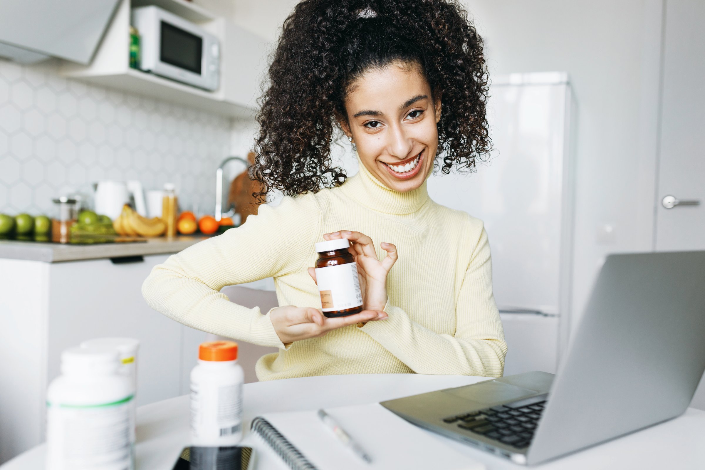 Young attractive woman of 20s advertising food supplement holding bottle in hands, recommending poly-unsaturated fatty acids or vitamin D3, sitting at kitchen table in front of laptop
