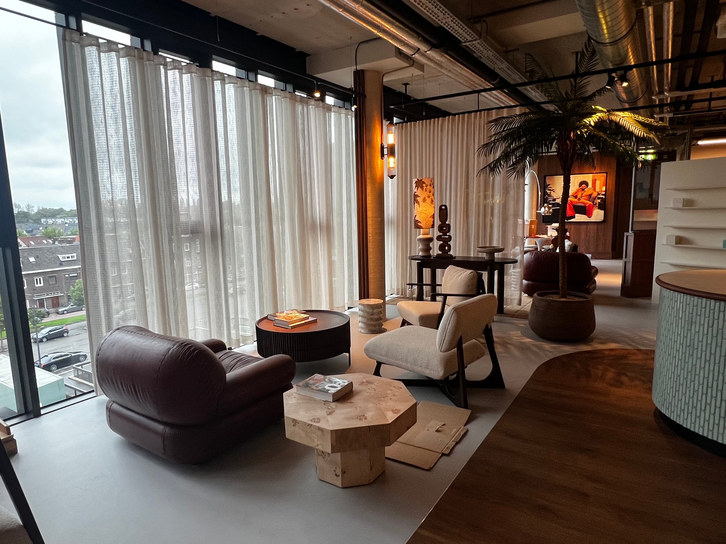 Modern lounge area with cushioned chairs, round tables, palm plant, and sheer curtains overlooking a cityscape.