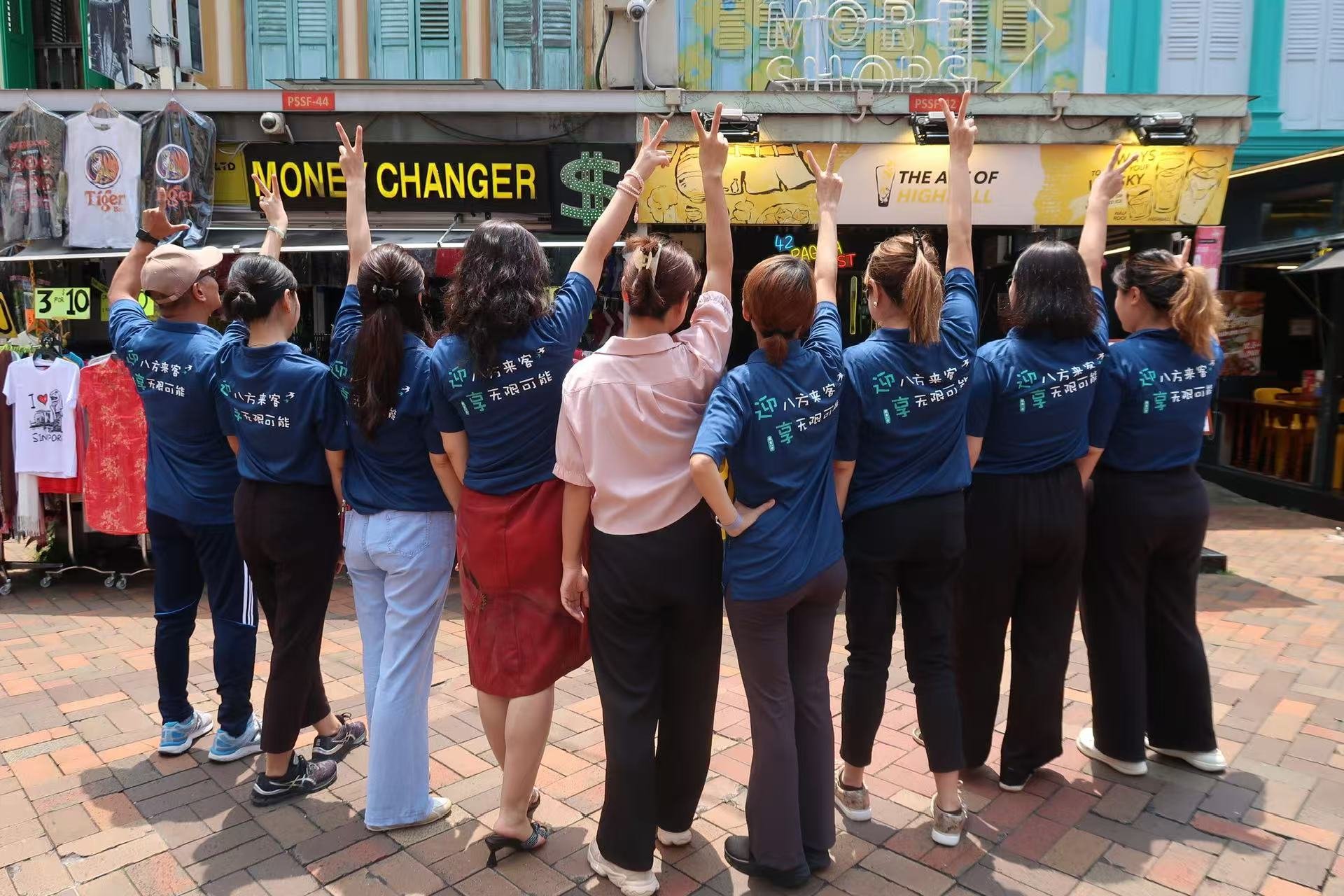 A group of people wearing blue shirts pose with their backs to the camera in an outdoor market area.