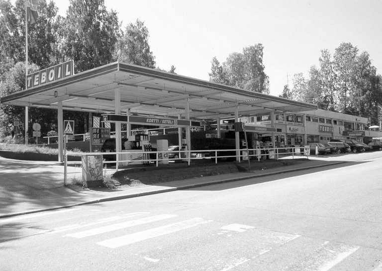 Black and white image of a Teboil gas station with several fuel pumps and parked cars, surrounded by trees.